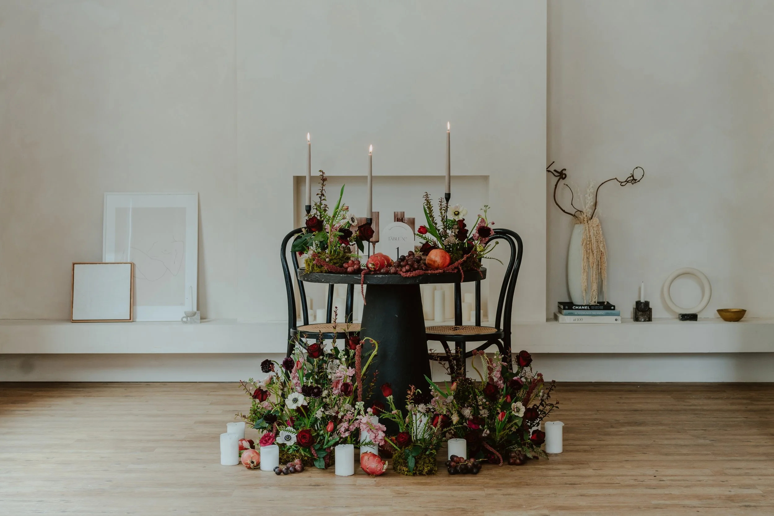 An interior scene with a round black table decorated with candles, flowers, and pomegranates, surrounded by two black chairs, with a floral arrangement on the floor, and various art pieces and books on a white shelf in the background.