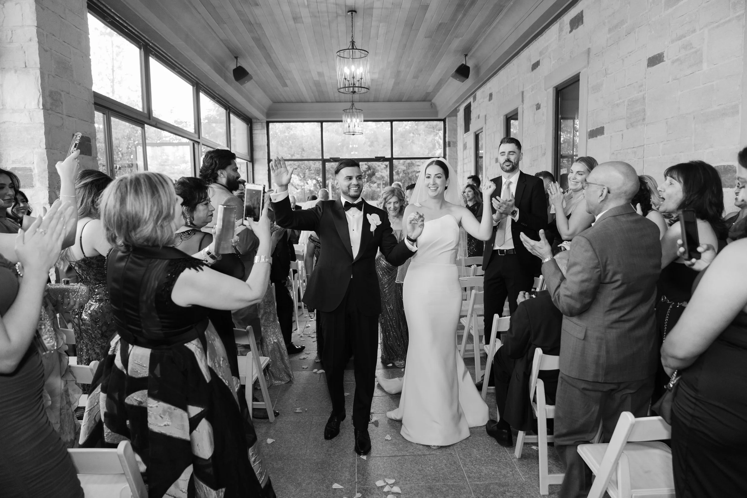 Black and white photo of a bride and groom walking hand in hand down the aisle at their wedding reception, surrounded by friends and family who are clapping and taking photos.