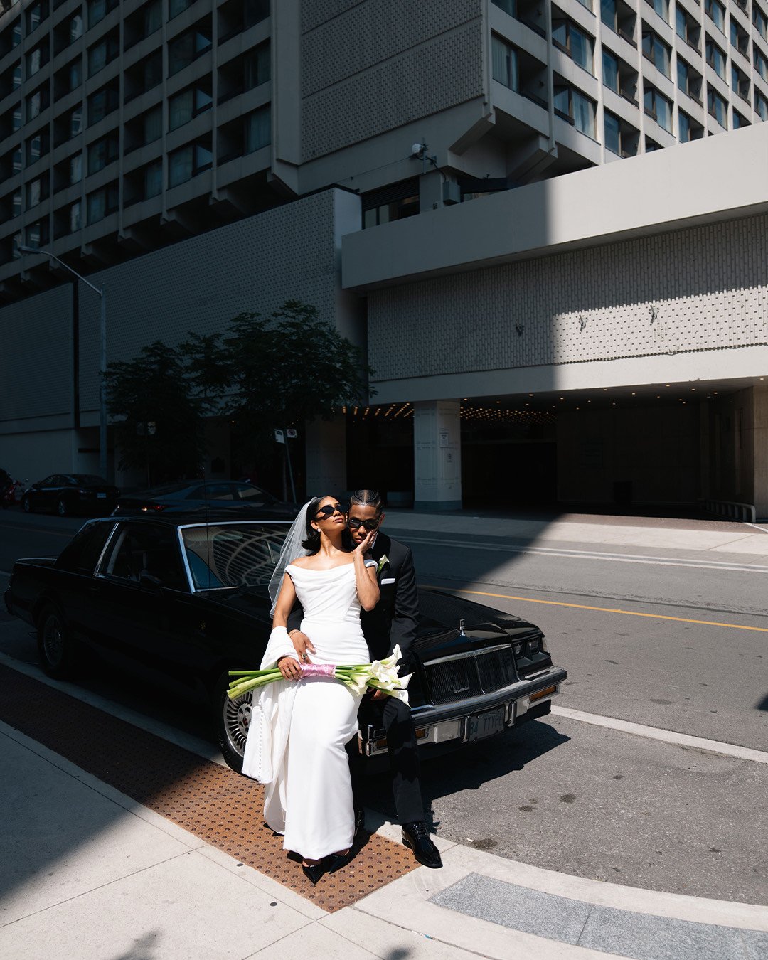 A newlywed couple sitting on the hood of a black vintage car in an urban setting, with a tall modern building in the background. The bride is wearing a white wedding dress and holding a bouquet of lilies, while the groom is dressed in a black suit and sunglasses. The scene is partly shaded and partly sunny.