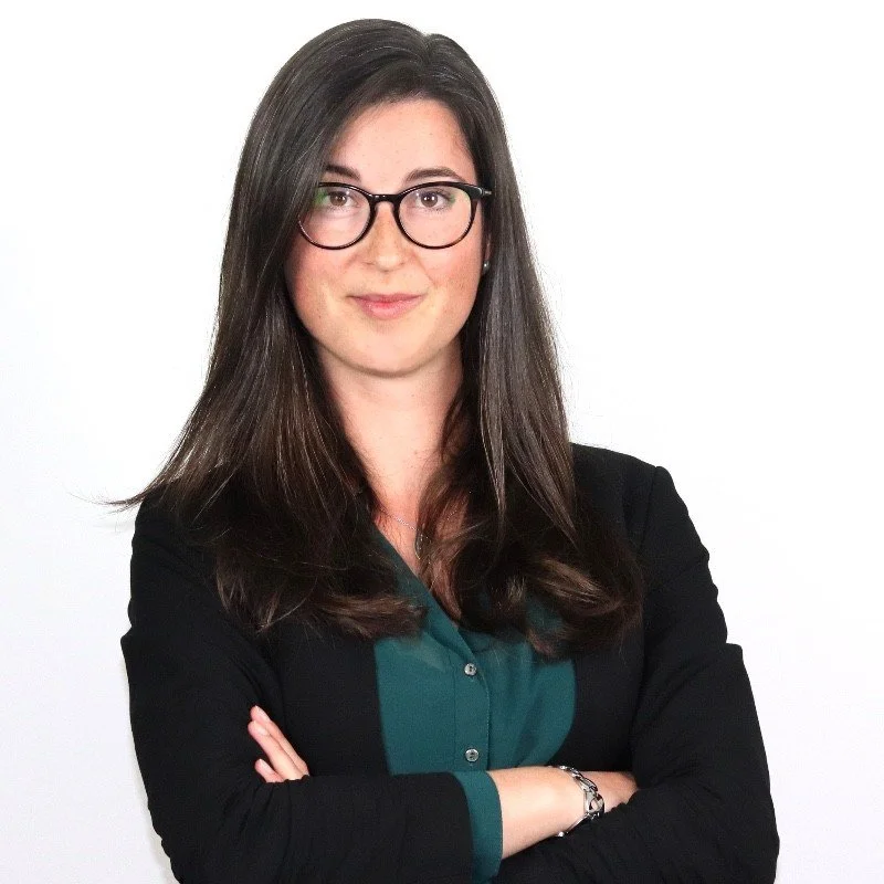 A woman with long brown hair, wearing glasses, a teal blouse, and a black blazer, standing with her arms crossed against a white background.