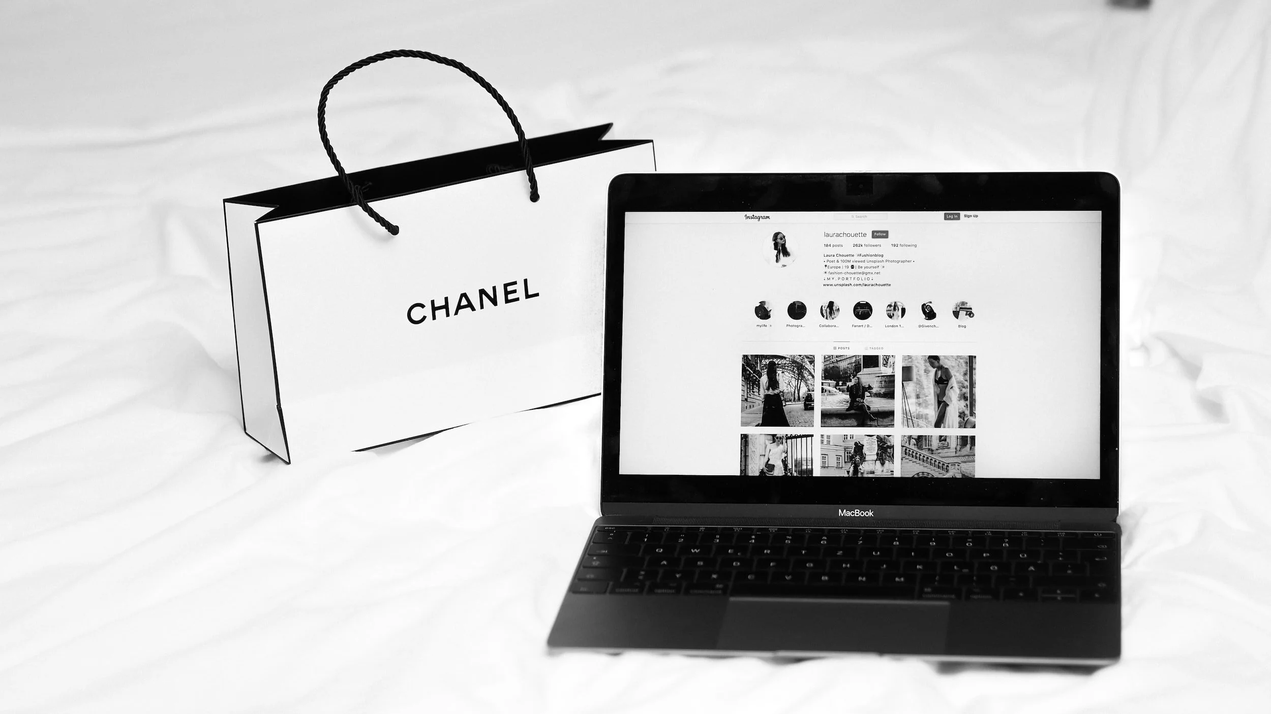 Black and white photo of a MacBook laptop displaying an Instagram profile, with a Chanel shopping bag beside it on a white fabric surface.