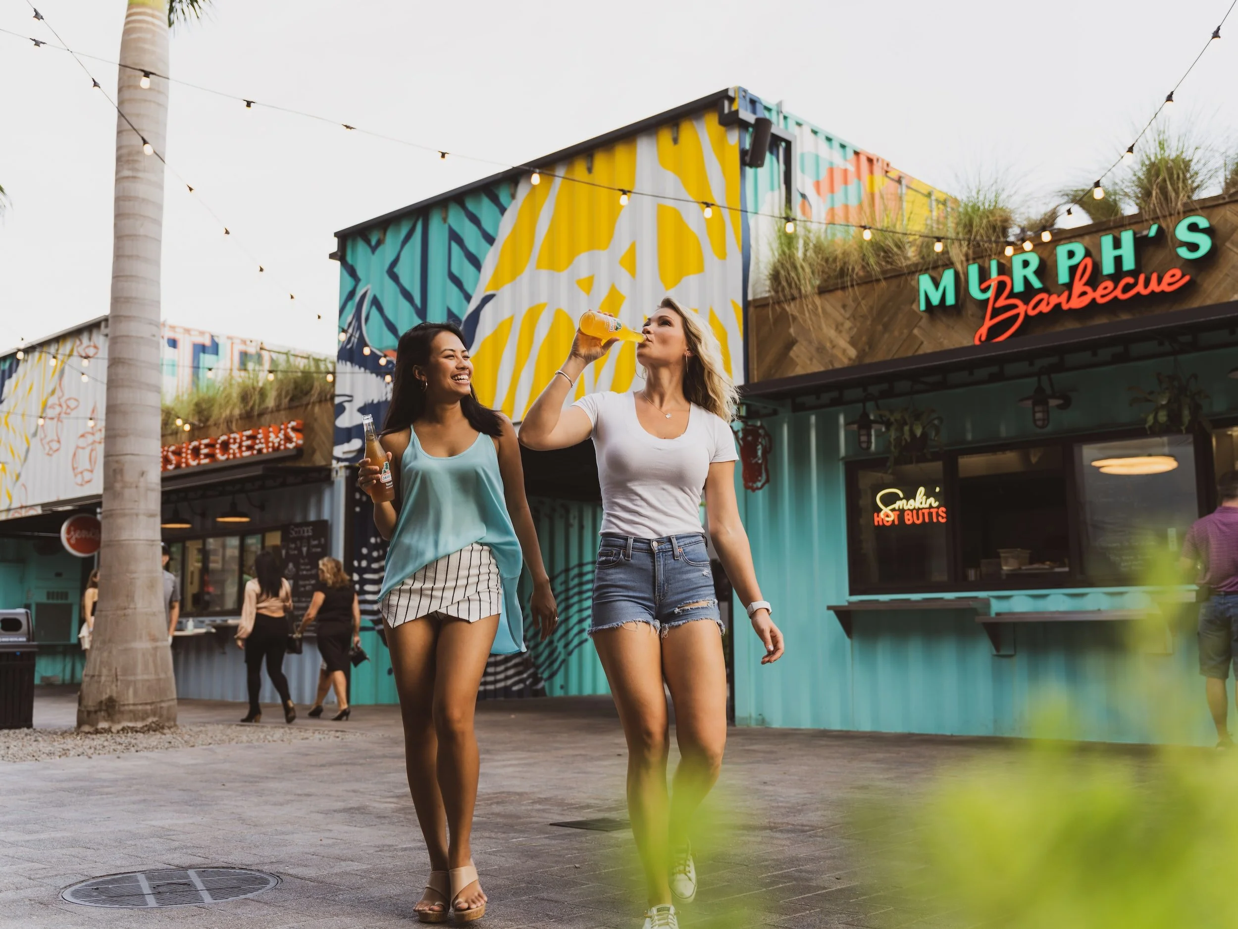 Two women walking and smiling outside a colorful bar or restaurant at dusk, one drinking a beverage and the other holding a bottle, with string lights overhead.