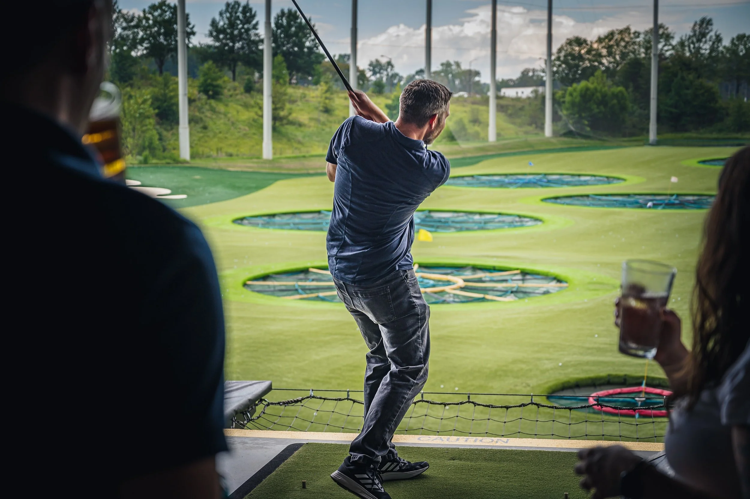 Man swinging a golf club at an indoor golf simulator, with two people watching and holding drinks in the foreground, and a view of a golf course projected on the screen.