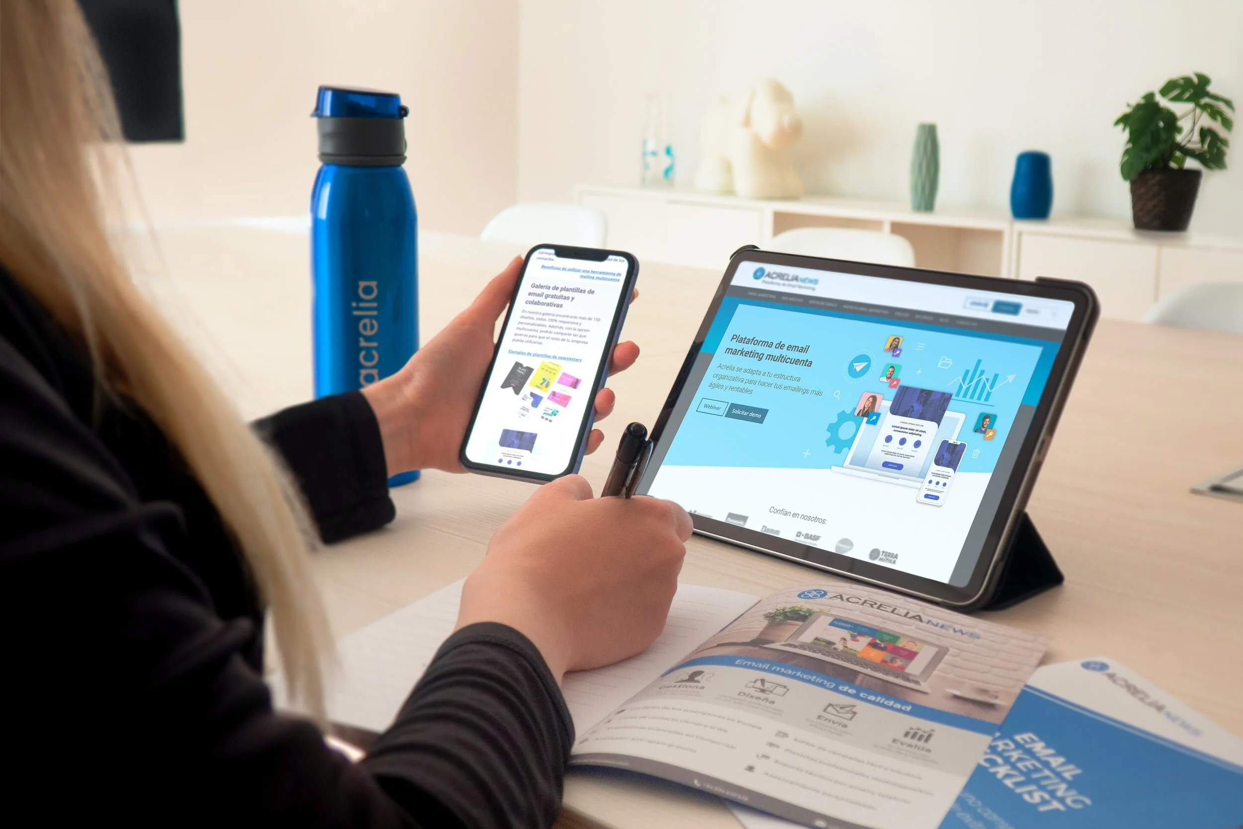 Person using a smartphone and tablet at a desk, with a blue water bottle and marketing materials nearby, in a modern office setting.
