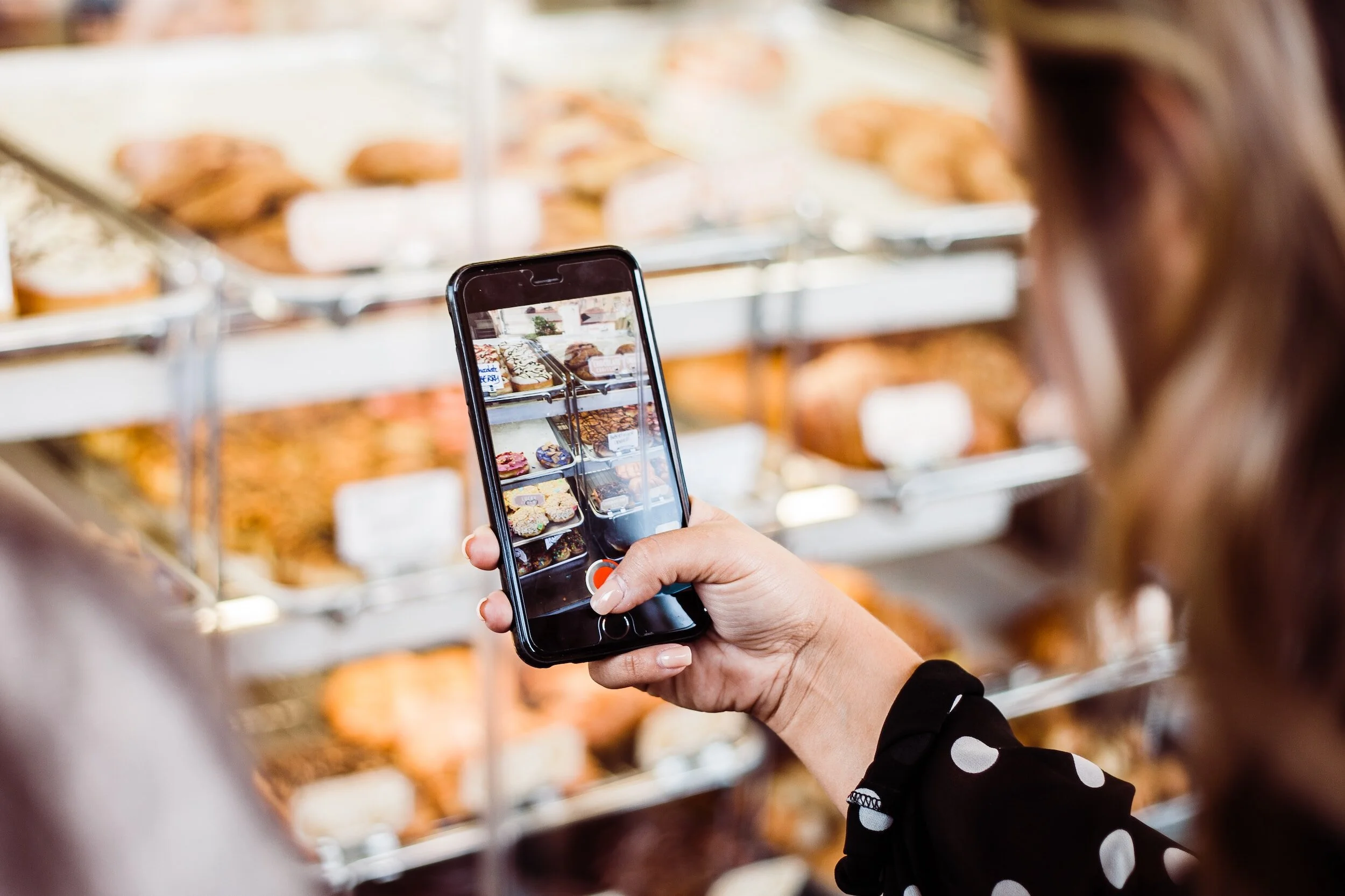 A woman taking a photo of baked goods on display in a bakery with her smartphone.