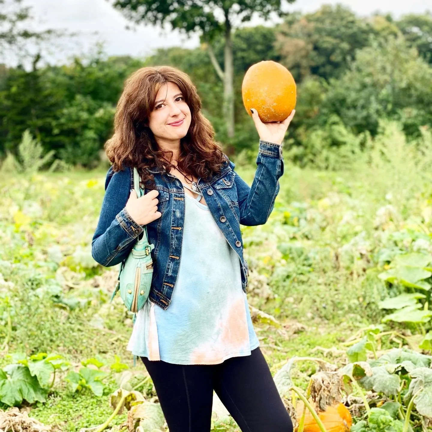 A young woman with curly brown hair, wearing a denim jacket and tie-dye dress, holding an orange pumpkin in an outdoor pumpkin patch.