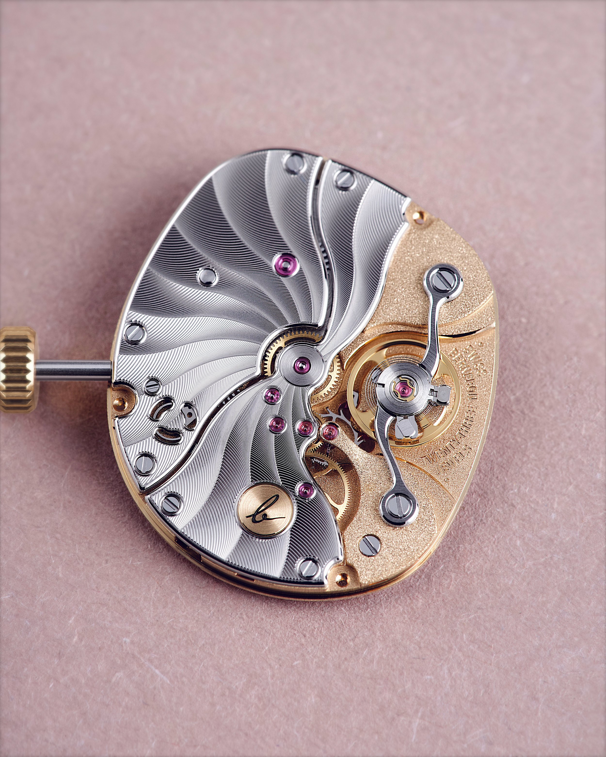 Close-up of a watch movement with intricate gears and polished metal components on a pink background.