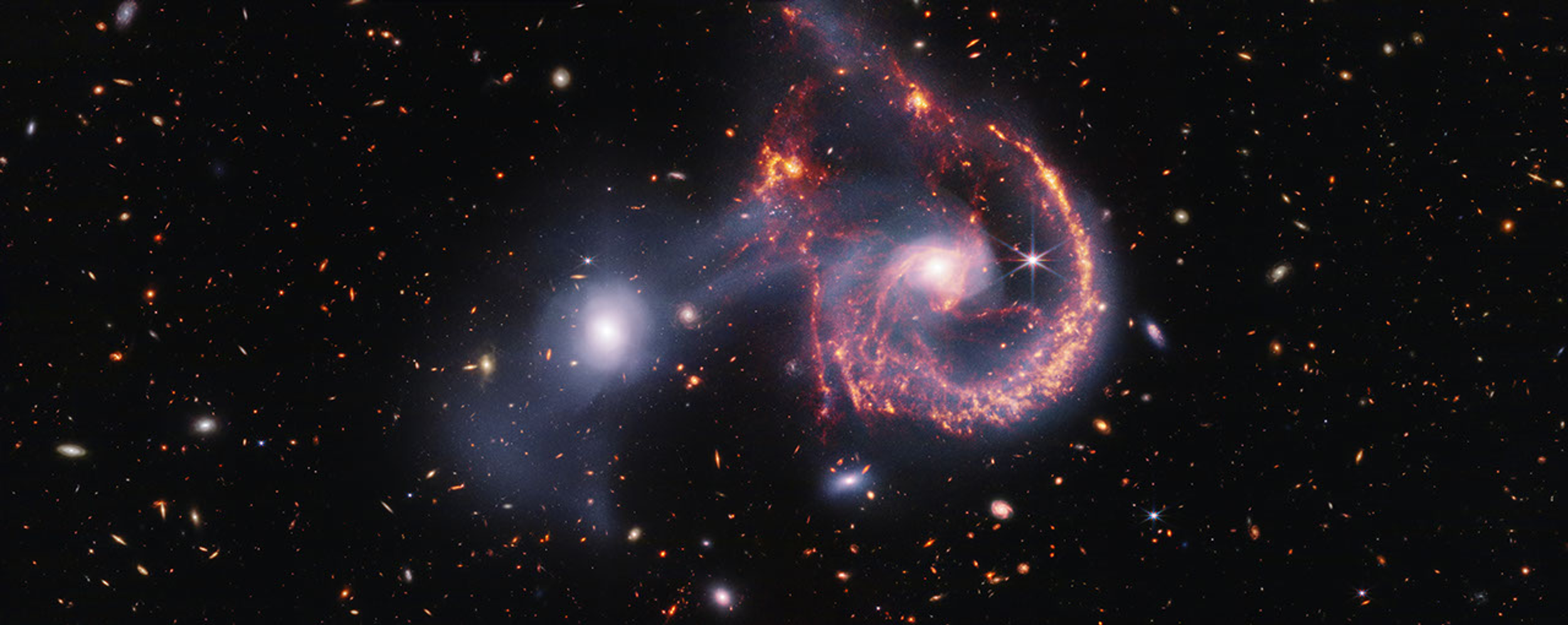 Las galaxias en interacción Arp 107, observadas por el telescopio James Webb. La imagen muestra estrellas y gas entre ambas galaxias, así como regiones de formación estelar resaltadas en tonos anaranjados y rojos.