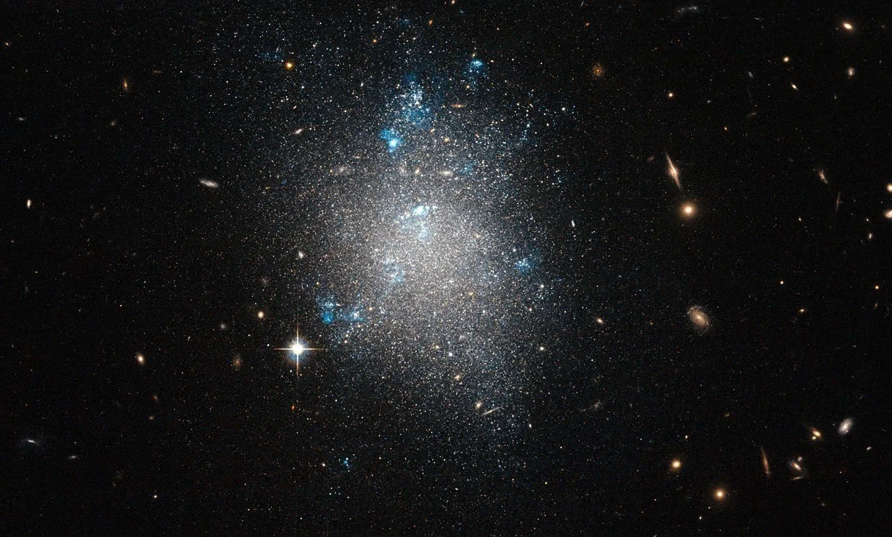 NGC 5477, una pequeña galaxia enana irregular del grupo de Messier 101, aparece salpicada de regiones brillantes donde nacen nuevas estrellas, visibles como nubes de hidrógeno incandescente.