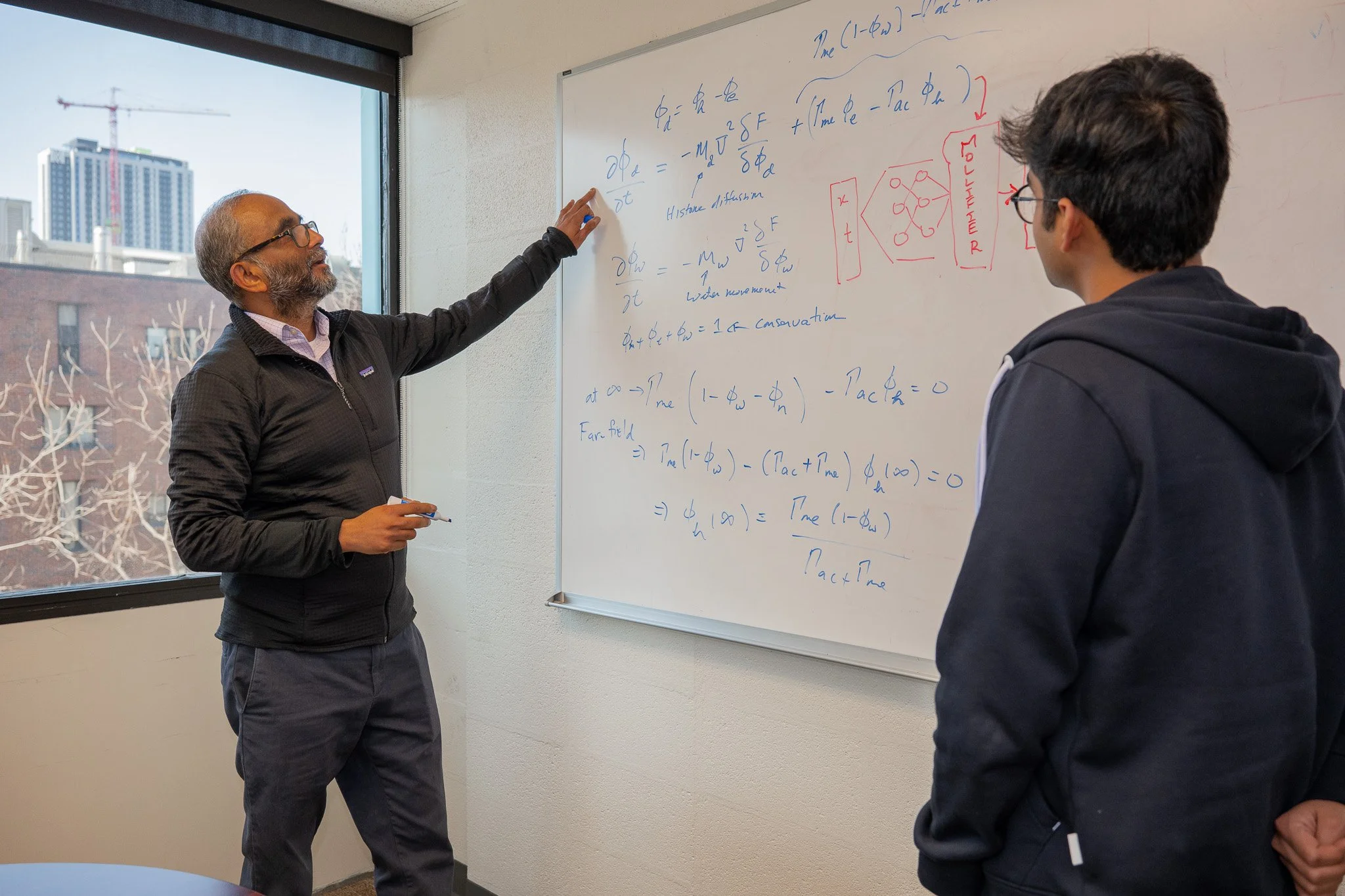 Vivek Shenoy (izquierda) explica en la pizarra los fundamentos matemáticos de las mollifier layers a Vinayak Vinayak (derecha), en una imagen que refleja el trabajo teórico detrás del avance en inteligencia artificial aplicada a ecuaciones complejas.