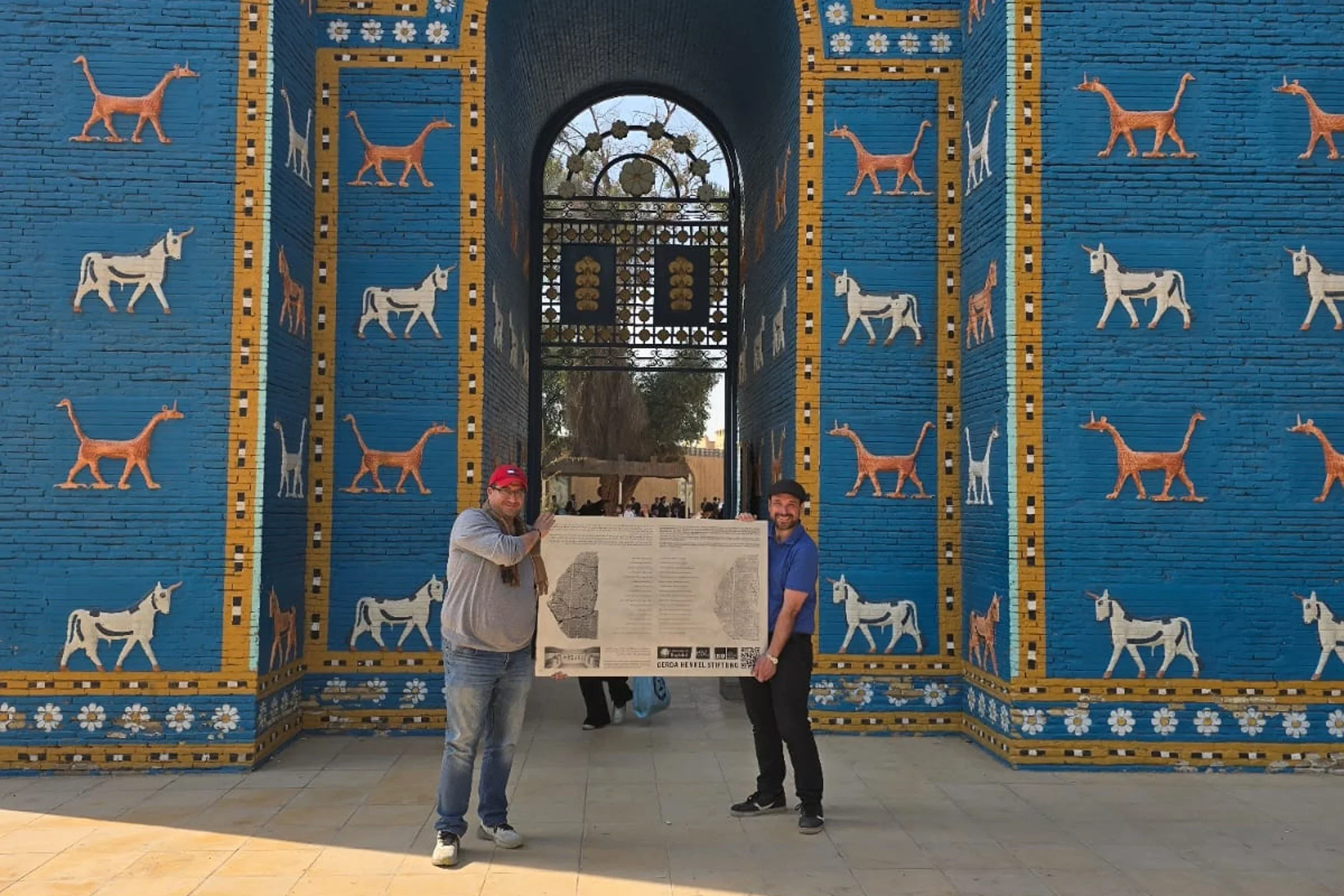 Enrique Jiménez y Anmar Fadhil, autores del estudio que reconstruyó un himno a Babilonia copiado por estudiantes, ante la réplica de la Puerta de Ishtar, a la entrada del yacimiento. Sostienen una placa con fragmentos del texto.