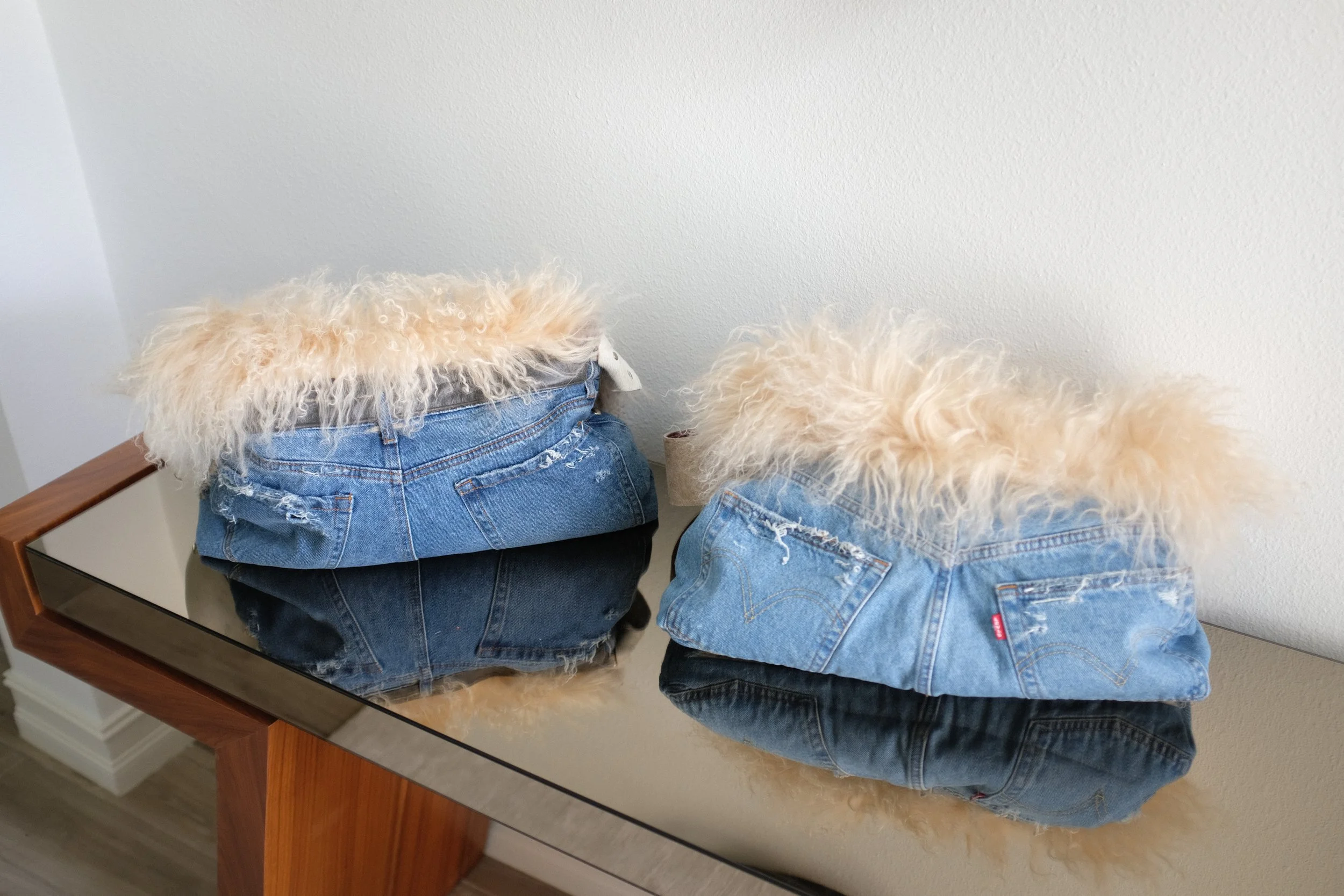 Two luxury handmade bags with real Mongolian sheep fur place on a wooden bench next to a mirror reflecting their images, in front of a plain white wall.