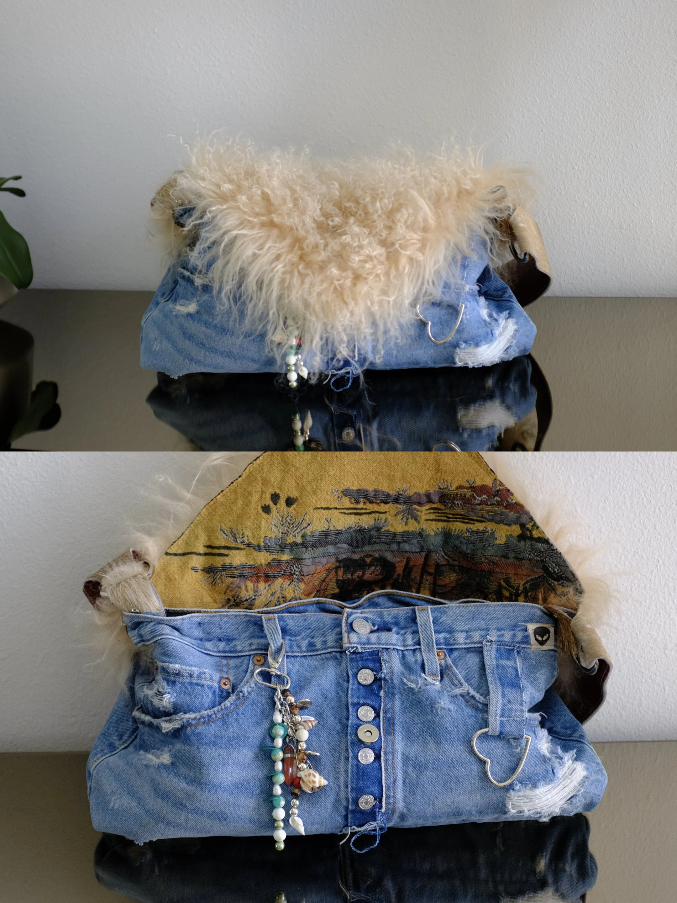 A fluffy beige dog lying on top of a pair of ripped blue jeans, which are placed over a black surface. The dog is partially covered by a blue denim jacket with a beaded necklace hanging from its neck. The background has a plain light-colored wall.