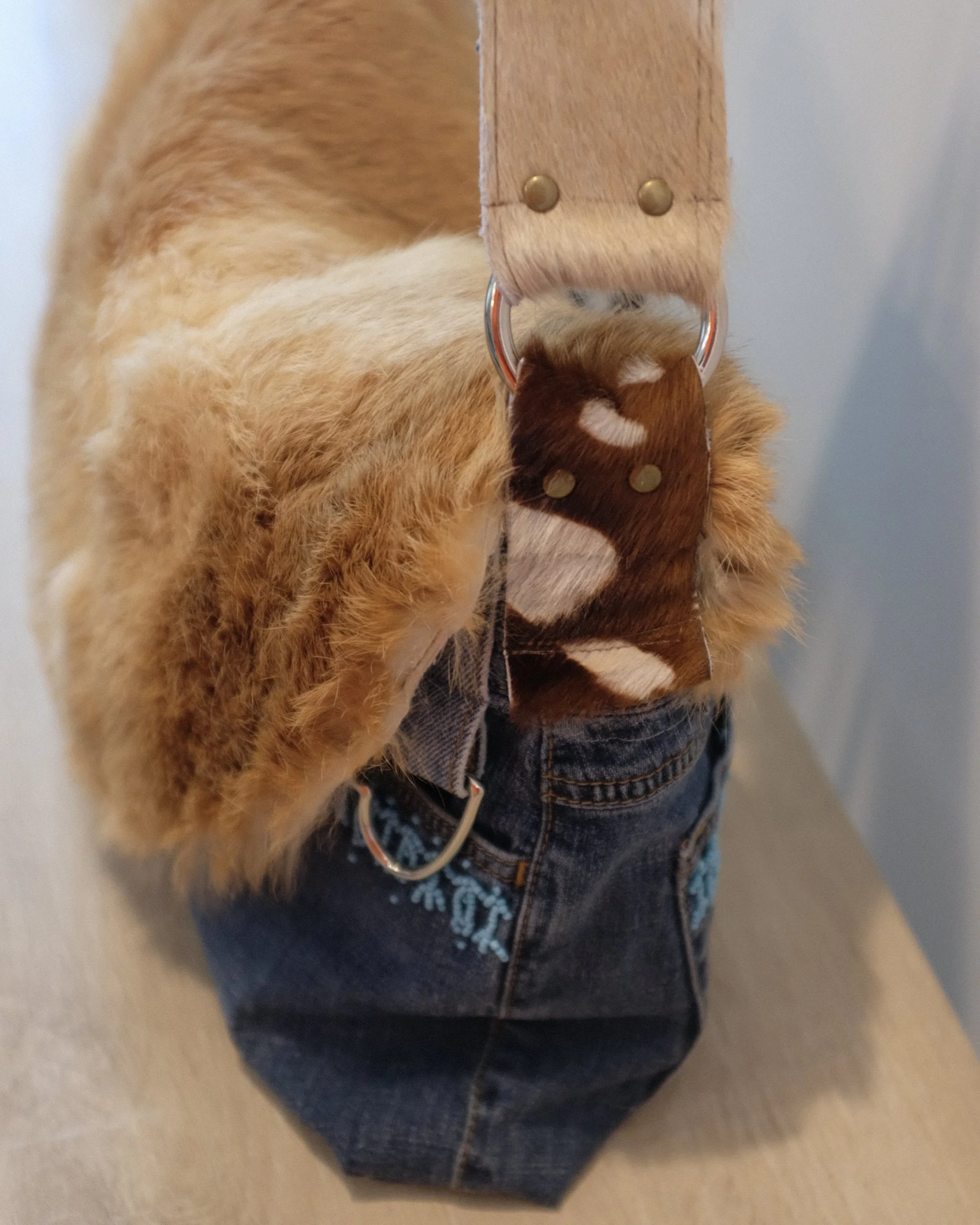 Closeup of handmade luxury denim handbag. The handbag is on a wooden table and features a cowhide shoulder strap and hand beading on the pockets.