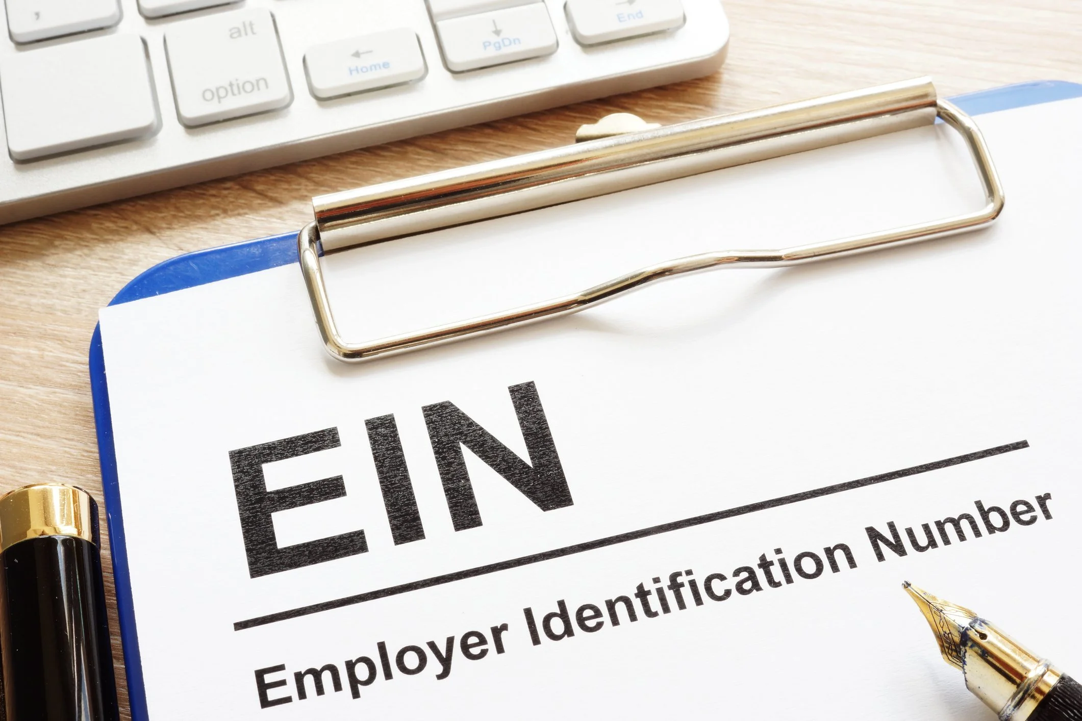 Close-up of a document titled 'EIN Employer Identification Number' on a clipboard, with a gold and black pen, a keyboard, and a yellow highlighter nearby on a wooden desk.