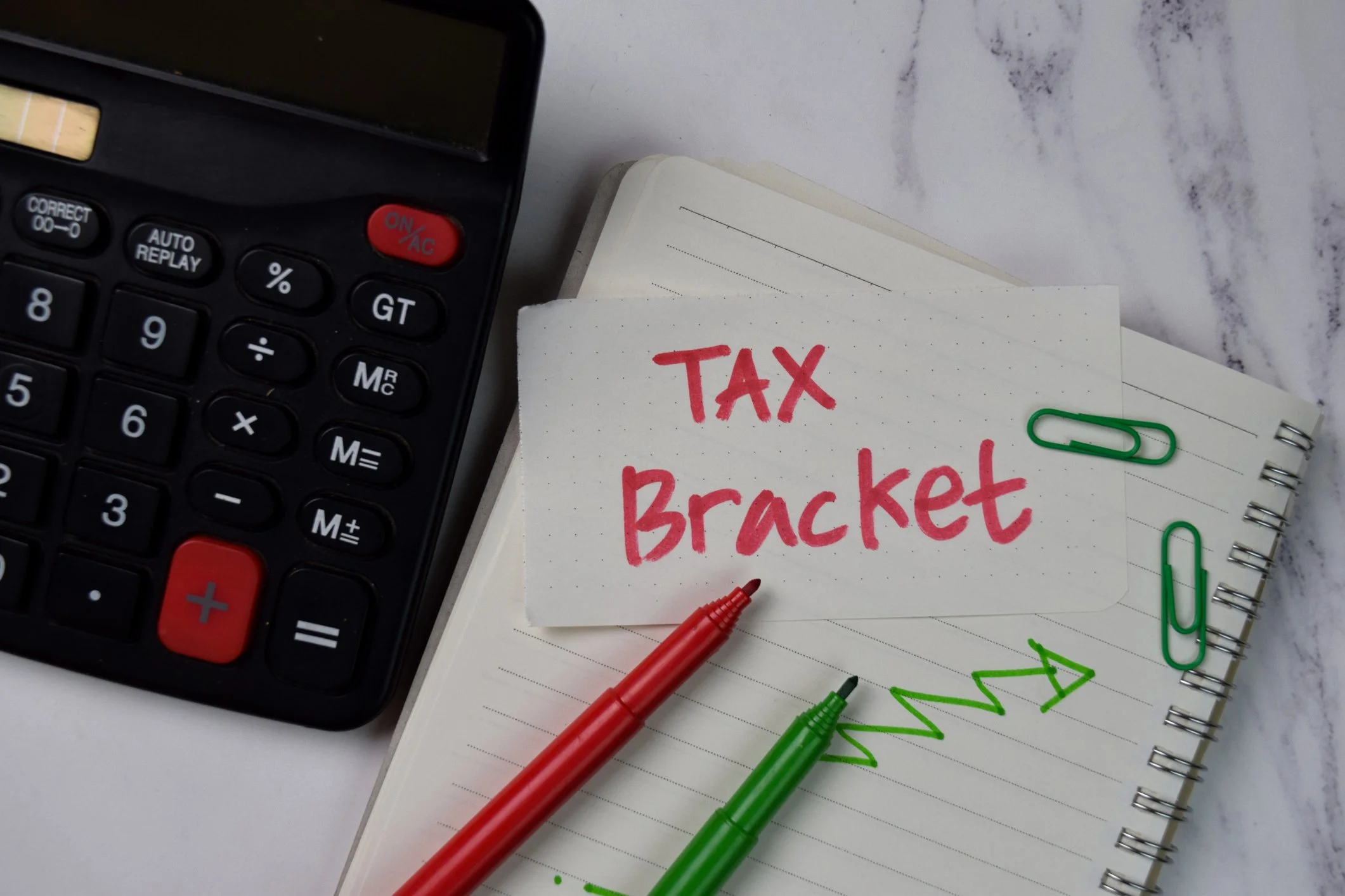 A calculator, a notebook with a green arrow drawing, a handwritten note saying 'Tax Bracket' in red, two pens (red and green), green paper clips, on a marble surface.