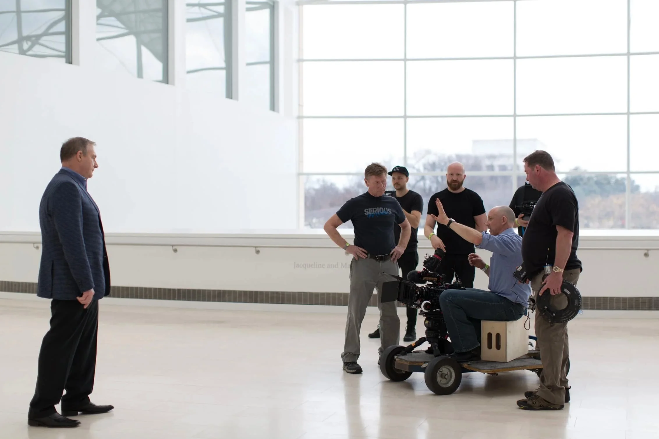 A group of six men on a film set inside a large, modern building with glass walls. One man is standing apart, facing the others who are gathered around a camera rig. The scene appears to be a filming or photoshoot session.