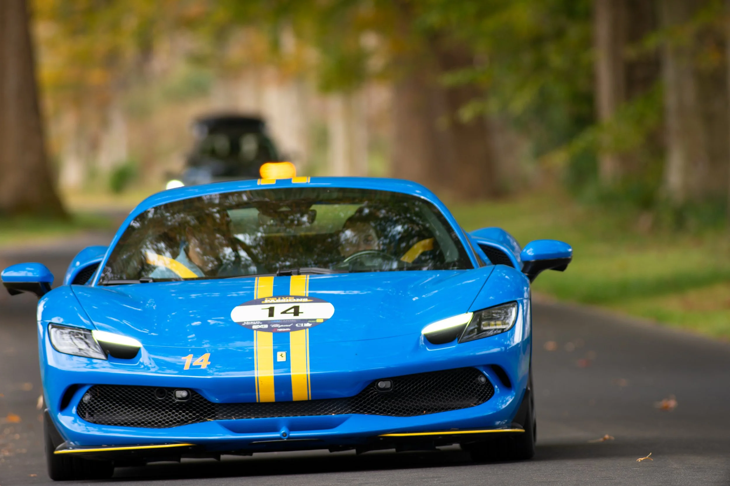 A blue race car with yellow racing stripes and number 14 on the front, driving on a tree-lined road during daytime.