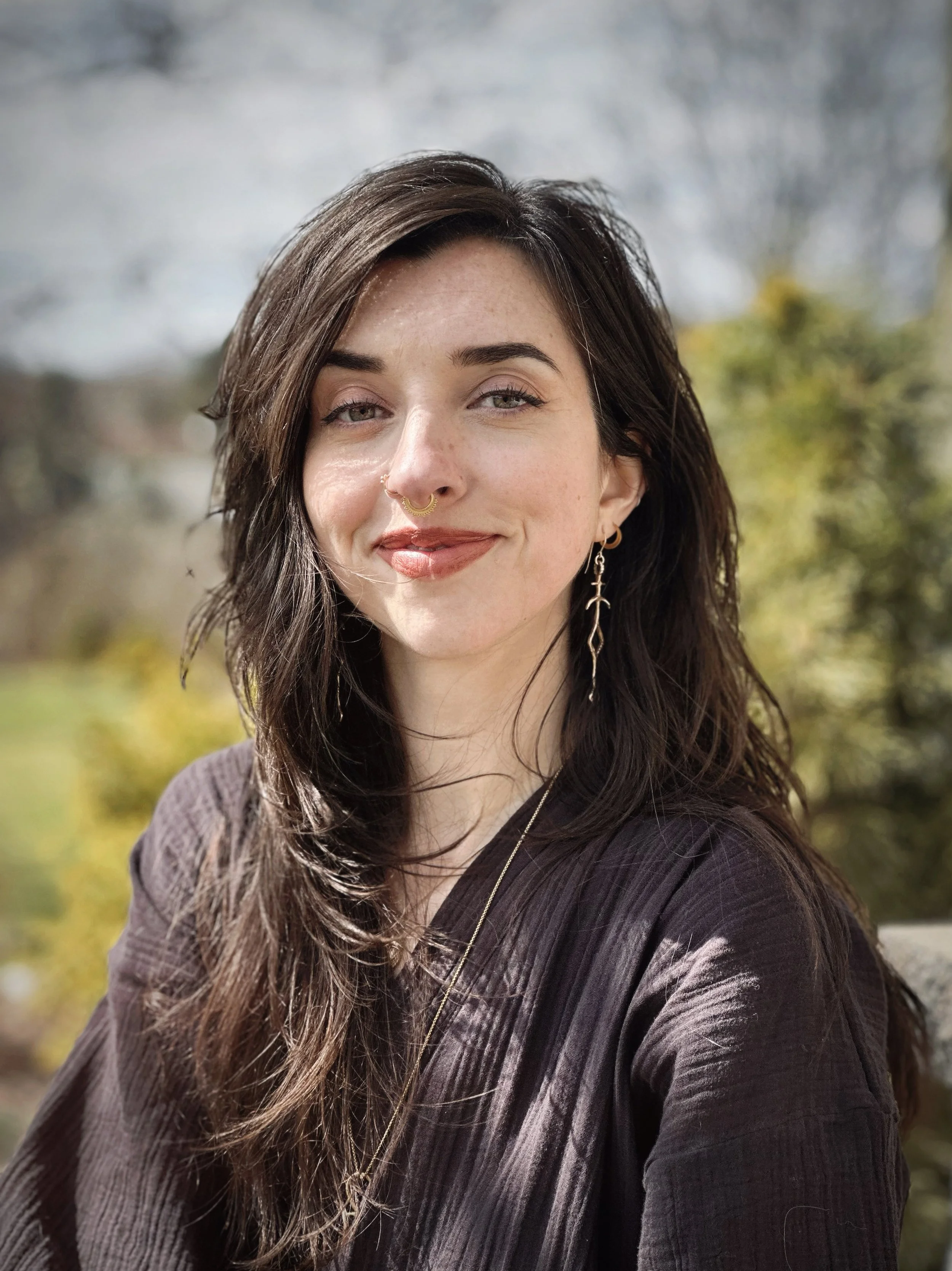 A woman with long dark hair and green eyes smiling outdoors, wearing gold jewelry and a dark top, with a blurred natural background.