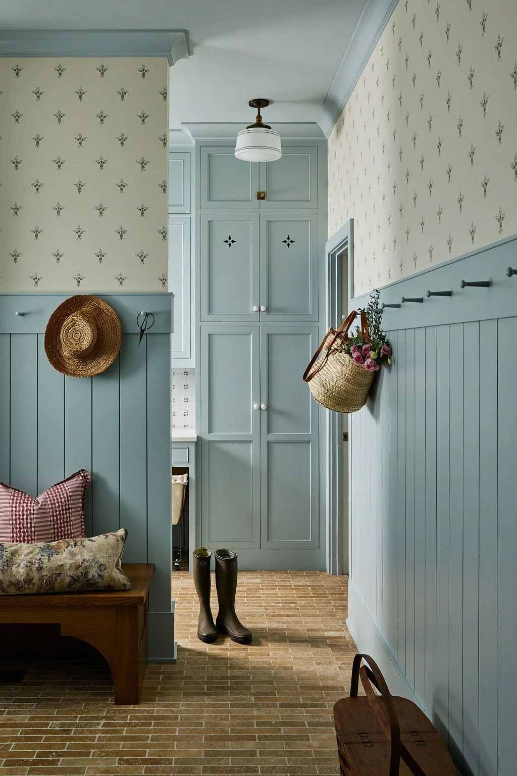 Mudroom with blue painted paneling, Chelsea Textiles wallpaper, built-in cabinets, a hanging straw hat, a pair of black rain boots, a basket with flowers, and a wooden bench with pillows, on a brick floor.