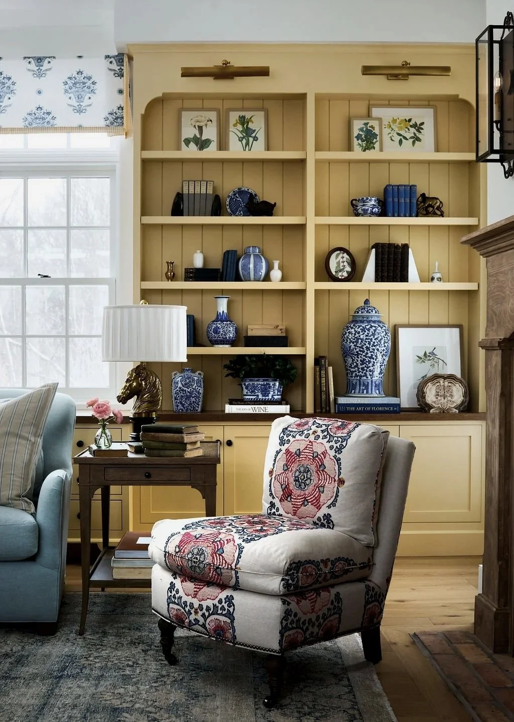 A cozy living room with yellow built-in bookshelf filled with blue and white vases, botanical prints, and books.  Embroidered floral armchair, light blue sofa , and a window with a patterned valance is in the background.