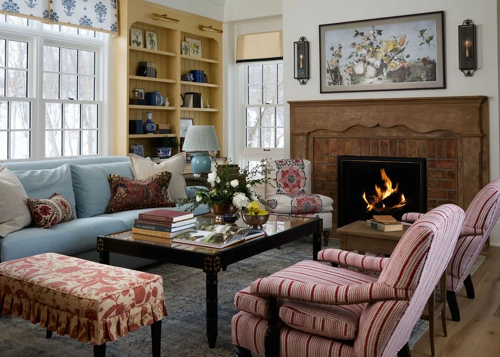Cozy living room with antique fireplace, striped armchairs, a light blue sofa with decorative pillows, a coffee table with books and flowers, a bookshelf with decor, large windows, and framed artwork.