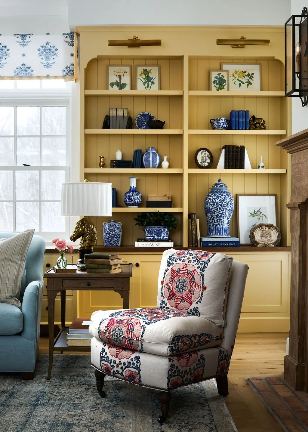 A cozy living room with yellow built-in bookshelf filled with blue and white vases, botanical prints, and books.  Embroidered floral armchair, light blue sofa , and a window with a patterned valance is in the background.