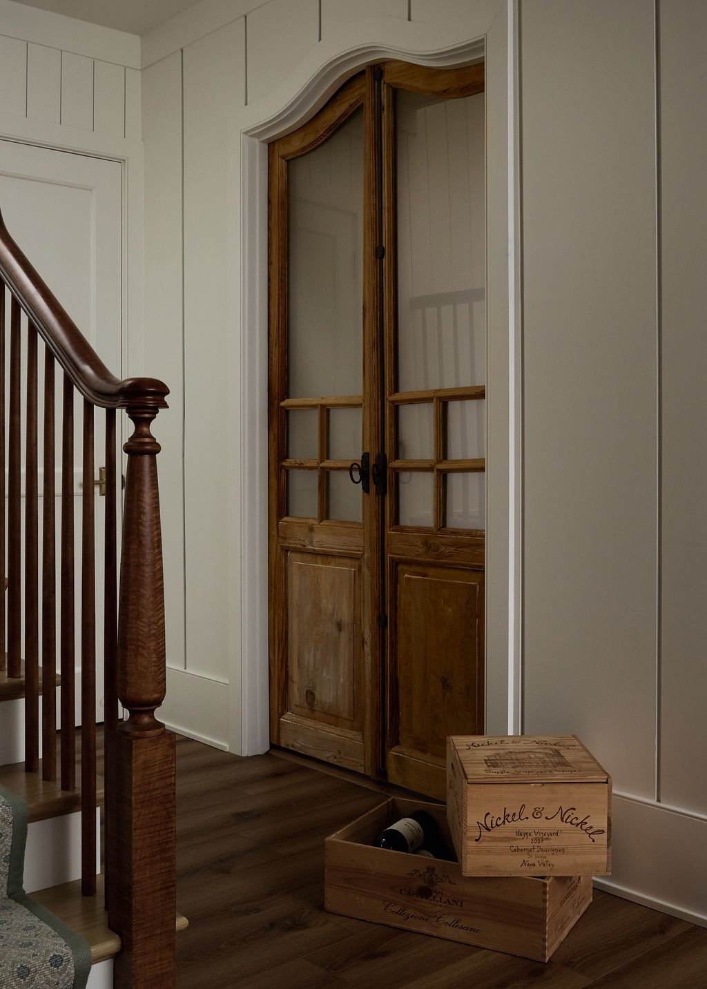 Antique doors with glass-panes in a hallway next to a staircase with wooden railing, on a hardwood floor, with a wooden wine box on the floor as entrance to wine cellar.