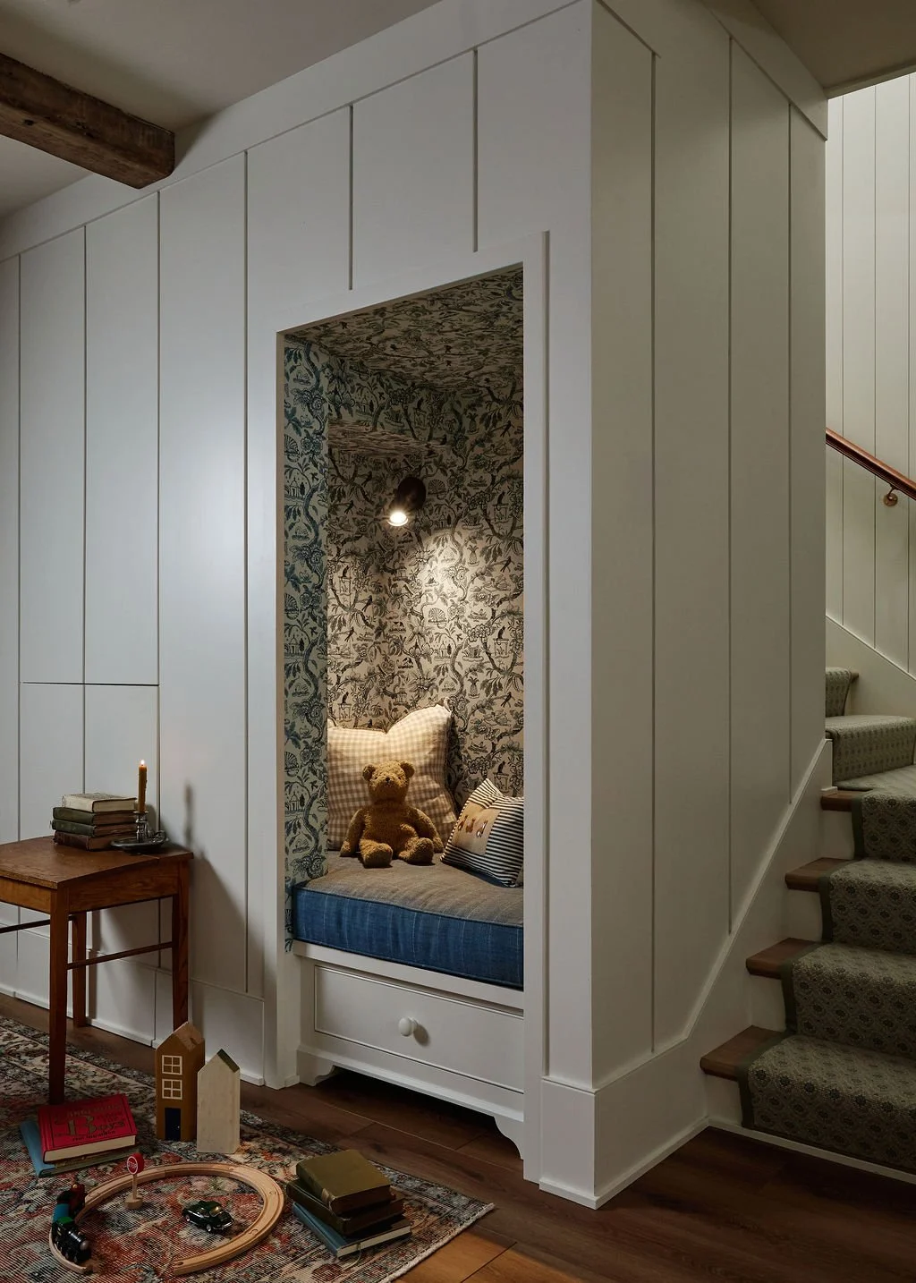 A cozy reading nook with a teddy bear, decorated wallpaper, and cushions, next to a staircase with patterned carpet. A small wooden side table with books and a candle is nearby.