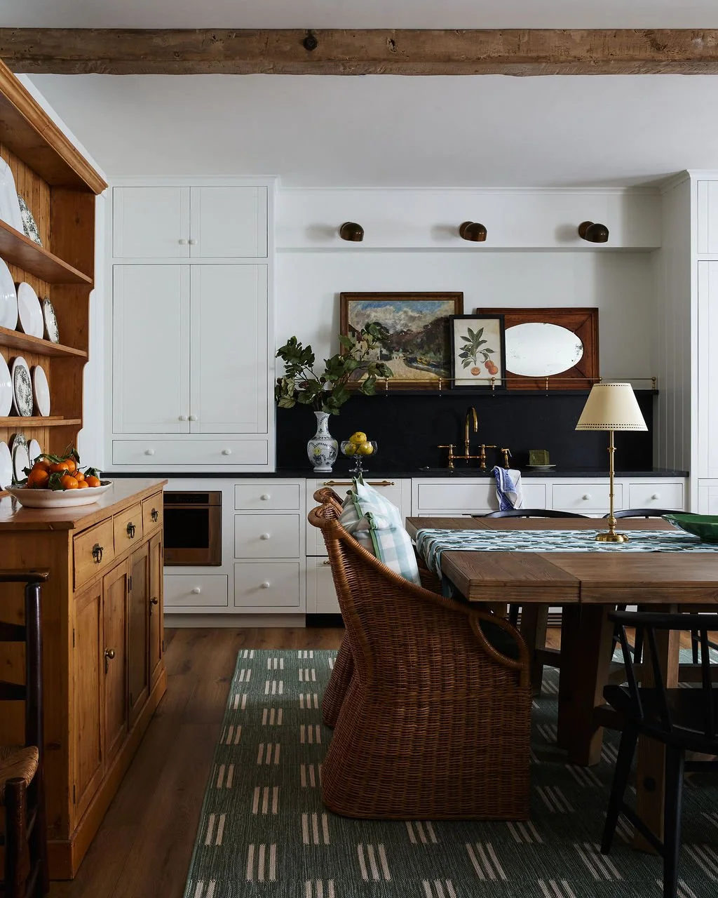 A kitchen and dining area with a wooden hutch on the left, white cabinets, a black backsplash, artwork on the wall, a table with a table runner, wicker chairs, a green and white patterned rug, and a lamp with a beige shade.