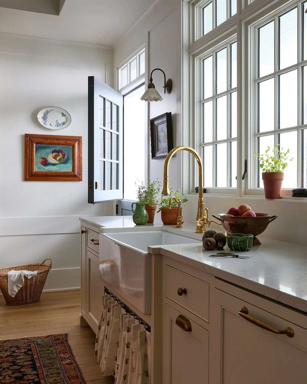 Bright kitchen with large windows, white cabinetry, blue dutch door and a brass faucet, decorated with potted plants and fruit.