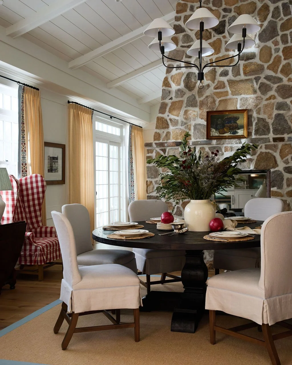 Living room with a round dining table, featuring a large white vase with greenery, surrounded by linen upholstered chairs. Double sided stone fireplace, gingham curtains, and a chandelier hanging from a white ceiling with exposed beams.