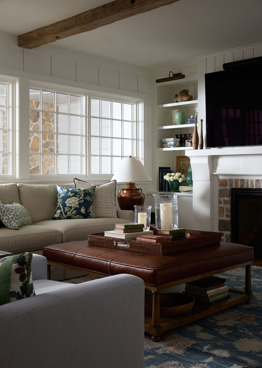 A cozy living room with a beige sofa decorated with patterned cushions, a brown leather ottoman with books and glass jars, a white armchair, and a fireplace with a mounted TV above it. There's a window letting in natural light and a white built-in bo