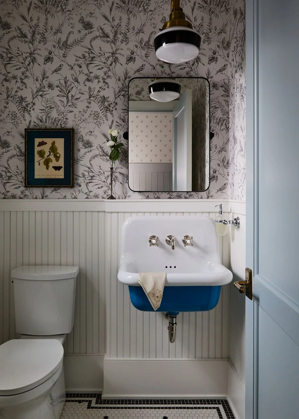 A small bathroom with a framed botanical print, a single rose in a vase, and a mirror reflecting a white door with toile wallpaper. Features include a toilet and a vintage-style blue and white utility sink.