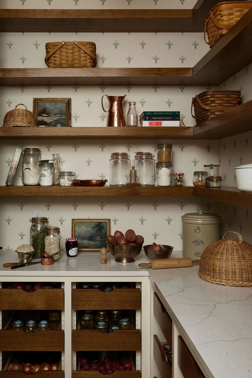 Kitchen pantry with open wooden shelves holding wicker baskets, jars of dry goods, books, a painting, and decorative items; a countertop with bowls of potatoes, jars, a rolling pin, and a vintage cookie jar.