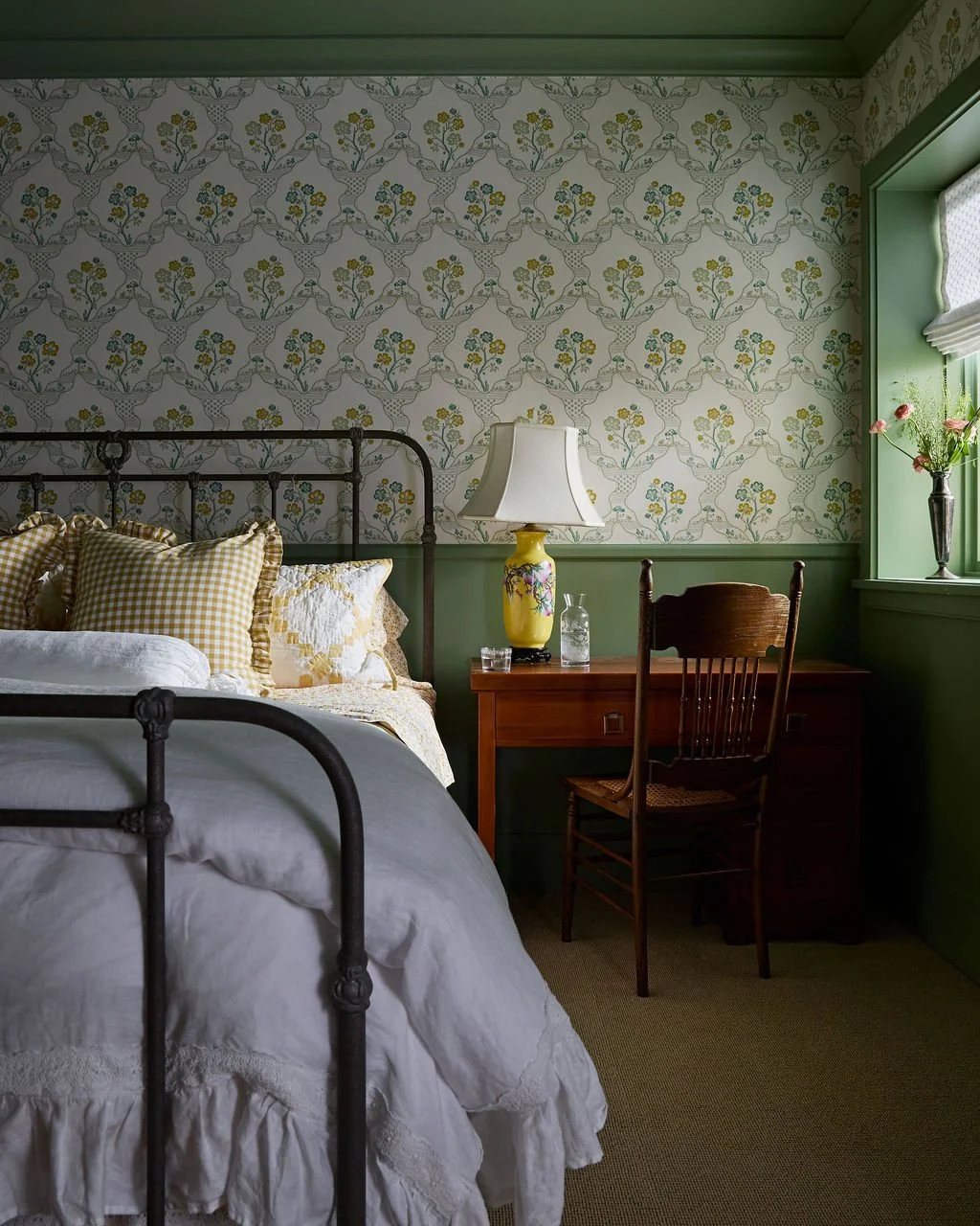 Cozy bedroom with a black metal bed frame, white bedding, and yellow gingham pillows. A wooden desk holds a yellow floral lamp, a glass pitcher, and a glass of water. A wooden chair is in front of the desk. A window with a green frame has a vase with