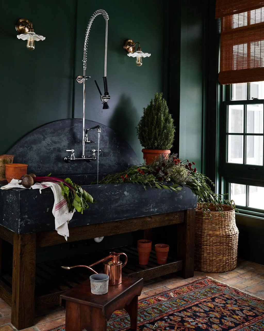 A solarium with a dark green wall, custom concrete sink, and vintage-style fixtures. There's a window with wooden blinds, a potted plant next to the sink, and various gardening tools and flowers on the counter and floor.