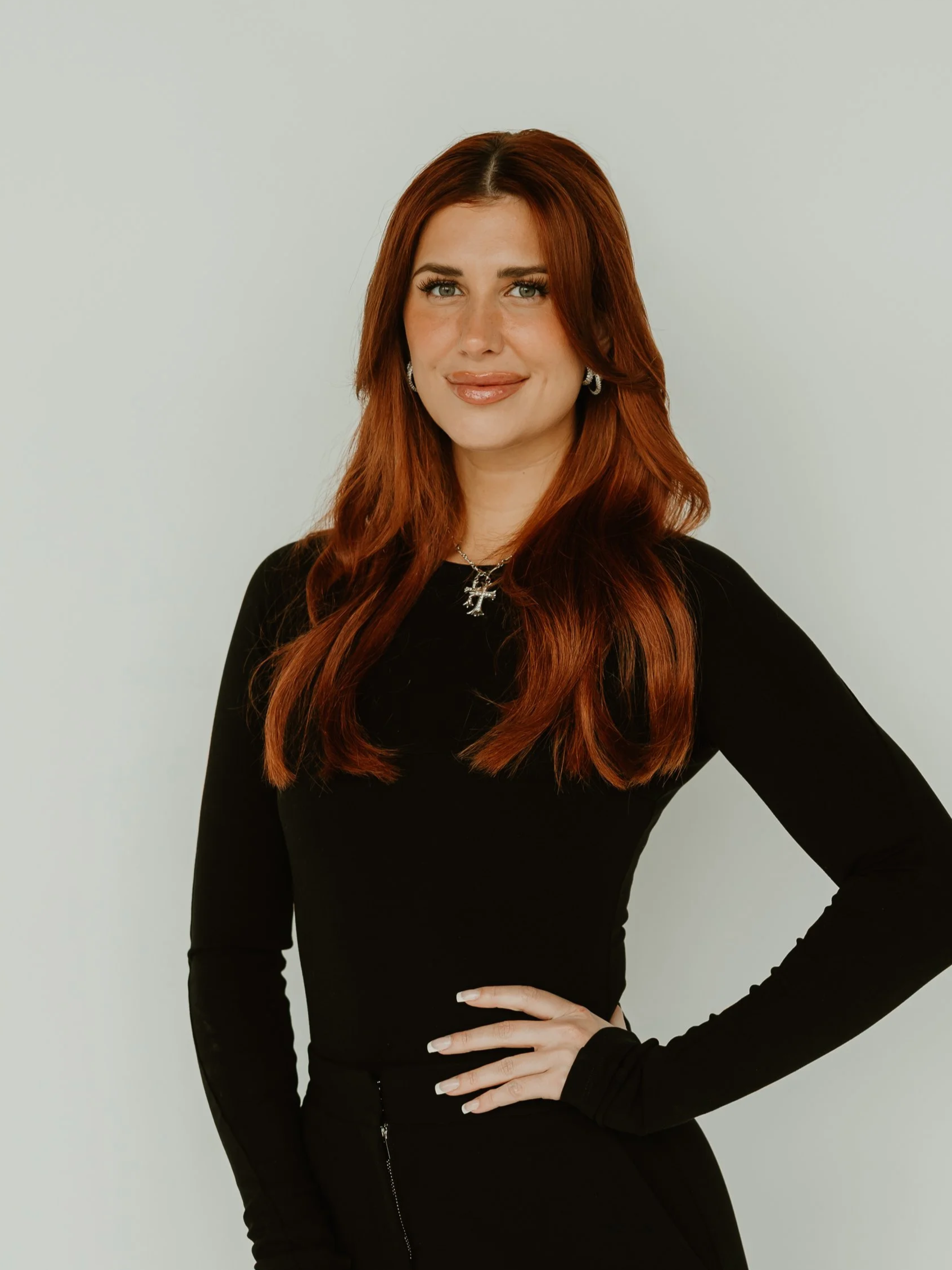 Woman with long blonde hair, wearing a black top and black pants, standing and smiling against a light background.