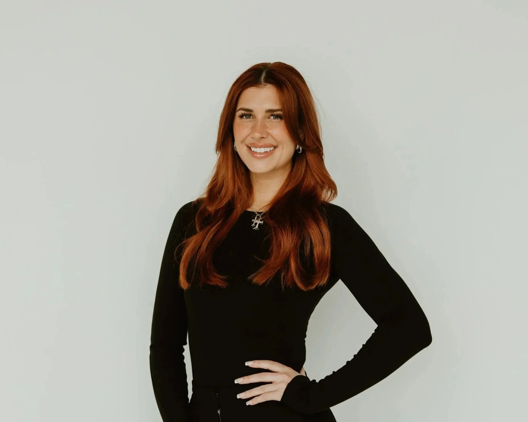 A woman with long red hair and blue eyes wearing a black long sleeve shirt, standing against a plain white wall, smiling with one hand on her hip.