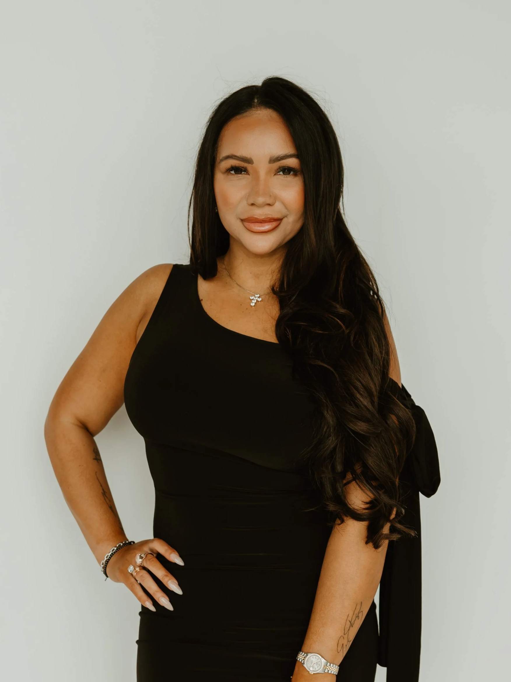 Woman with long dark hair in a black off-shoulder dress, smiling against a plain white background.
