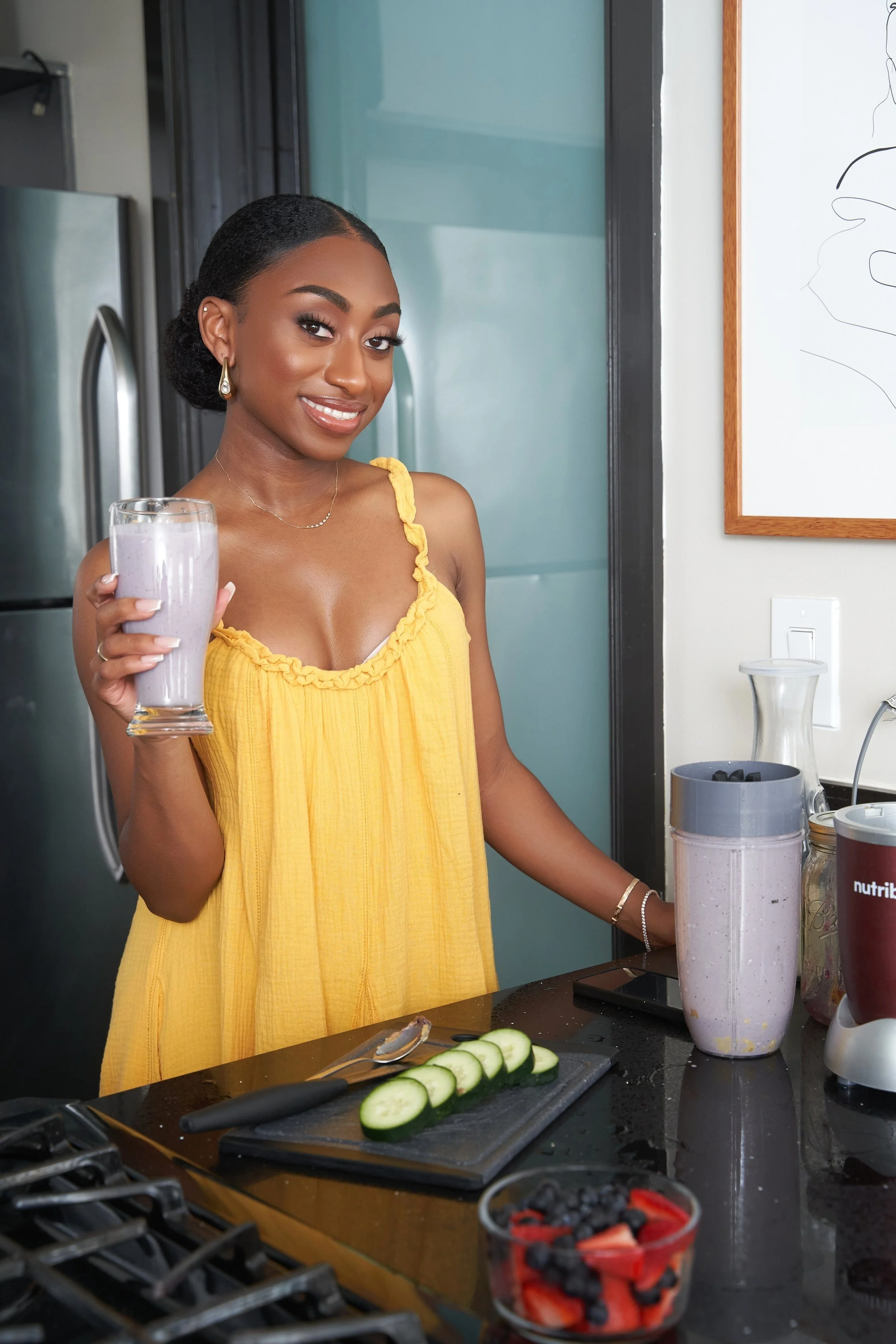 A smiling woman in a yellow dress holds a glass of purple smoothie in a modern kitchen with a black countertop, sliced cucumbers, fresh strawberries and blueberries, a blender, and other kitchen appliances.