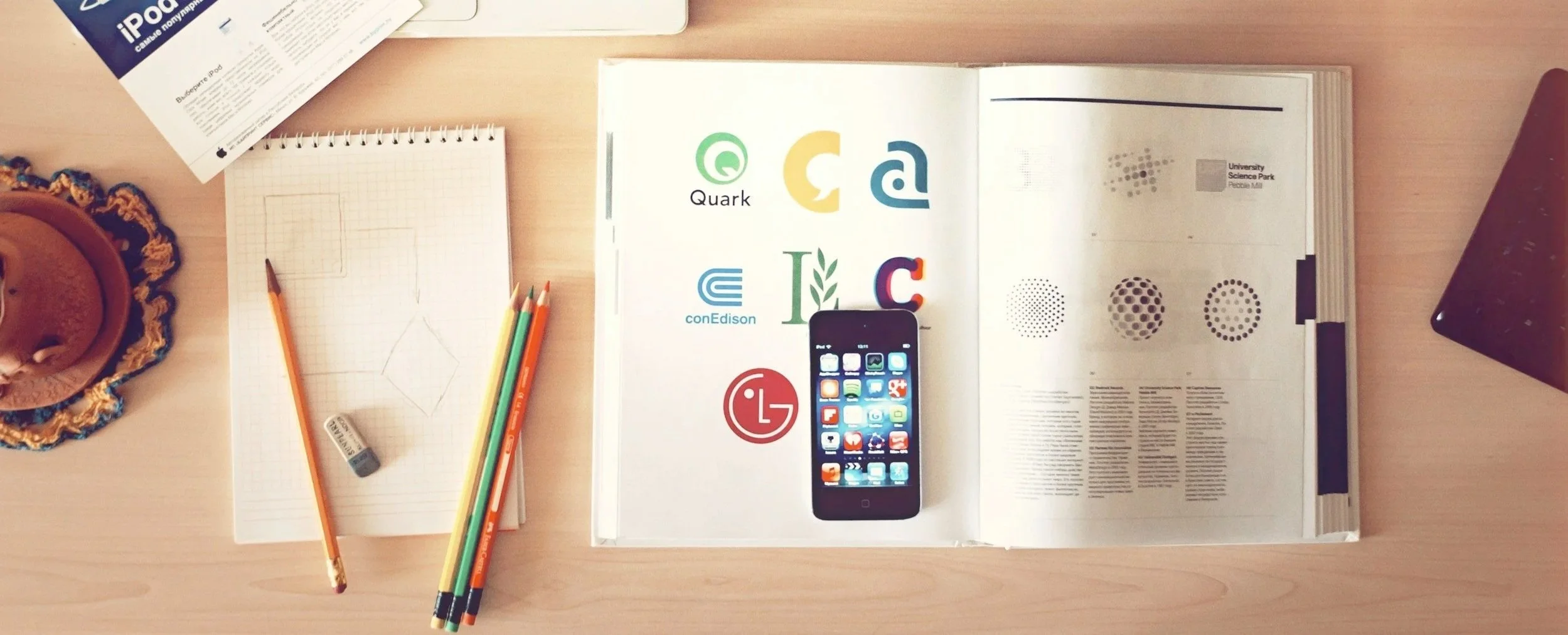 Top view of a workspace on a wooden desk with color pencils, a notebook with geometric sketches, an open magazine showing logos and images, a smartphone, and a small brown object on a decorative cloth.