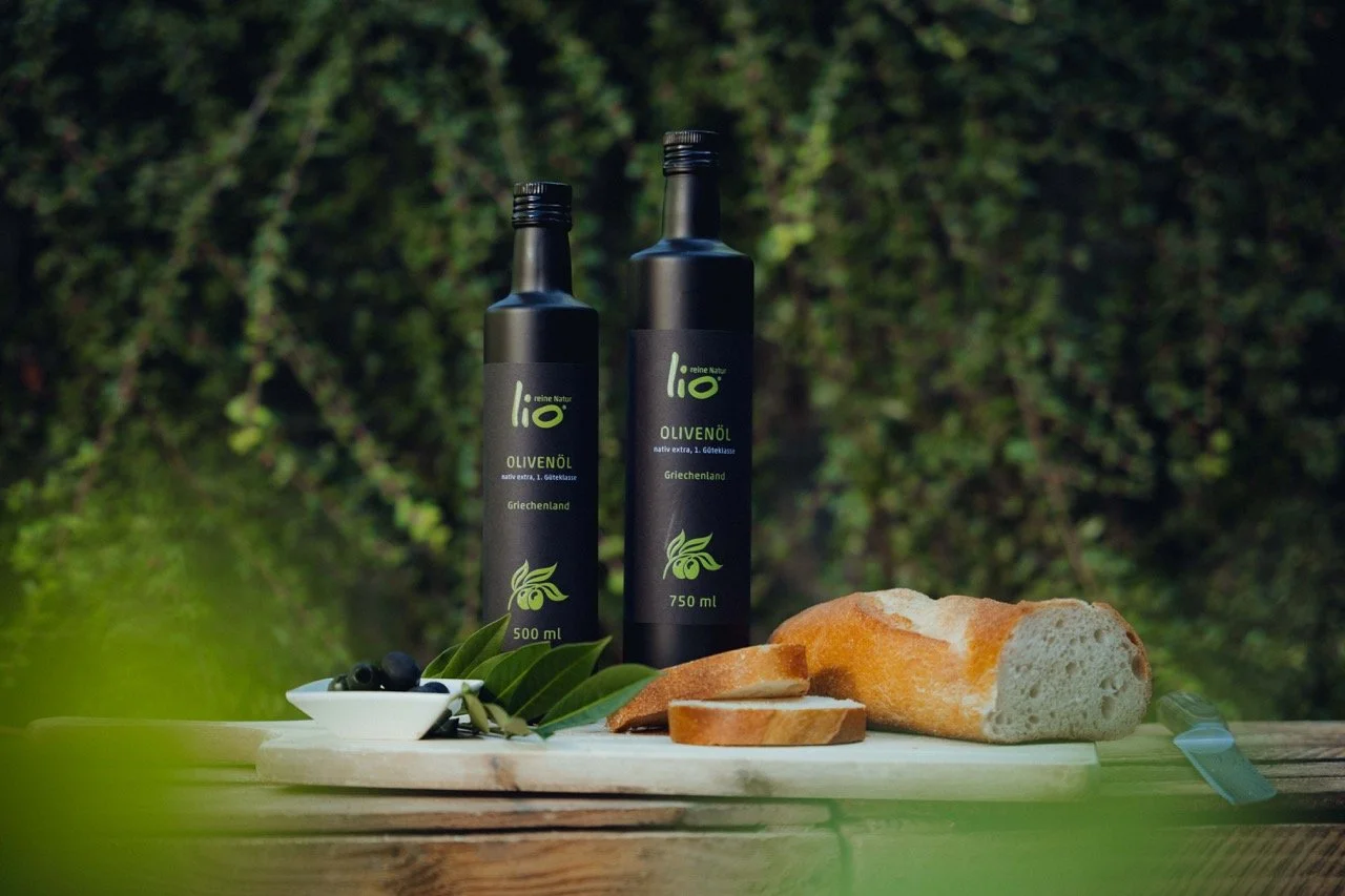 Two bottles of Greek olive oil, a loaf of bread, sliced, and some olives and green leaves on a white cutting board outdoors with green foliage background.