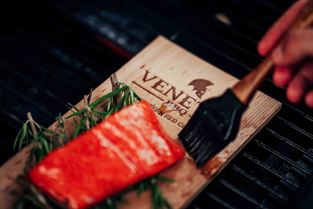 Brush applying glaze to a piece of salmon on a wooden board with sprigs of rosemary, on a black grill surface.