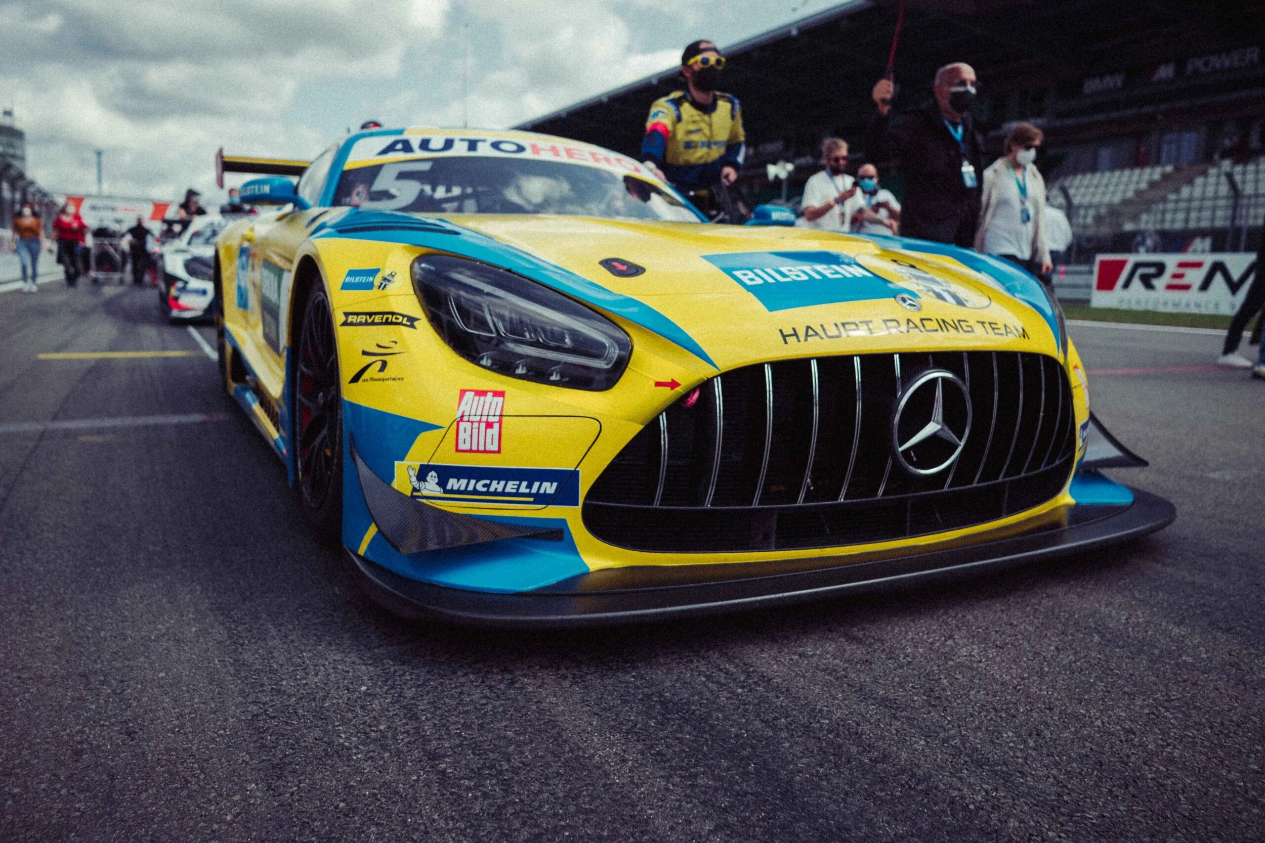A yellow and blue Mercedes-AMG race car on a race track with people in the background.