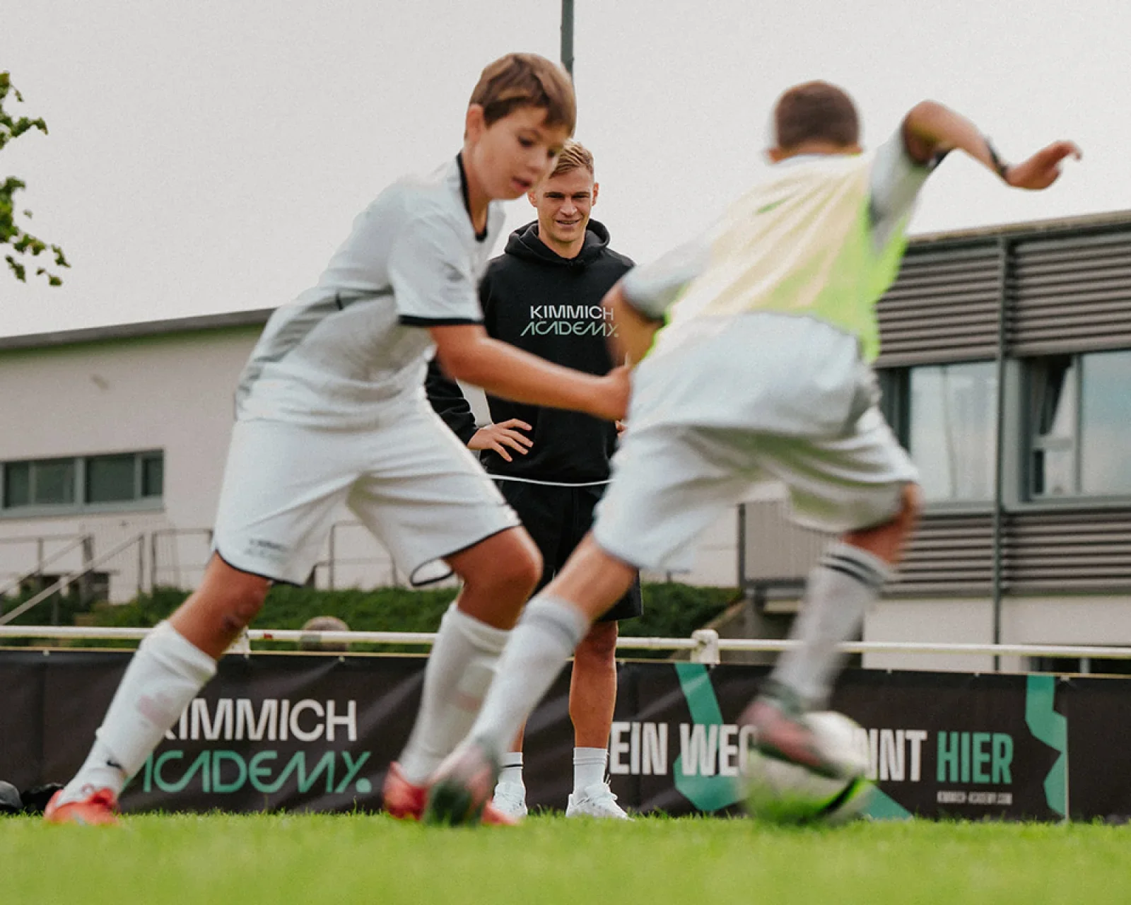 Two young boys playing soccer with an adult coach or referee watching on outdoors on a cloudy day.