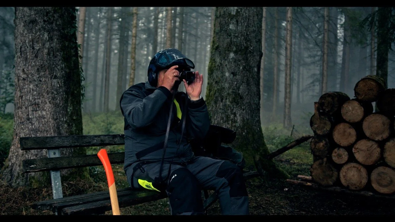 A person sitting on a park bench in a forest, wearing a black helmet, black jacket, and sunglasses, taking a photo with a camera. There are logs stacked to the right and a small axe with an orange handle leaning against the bench.
