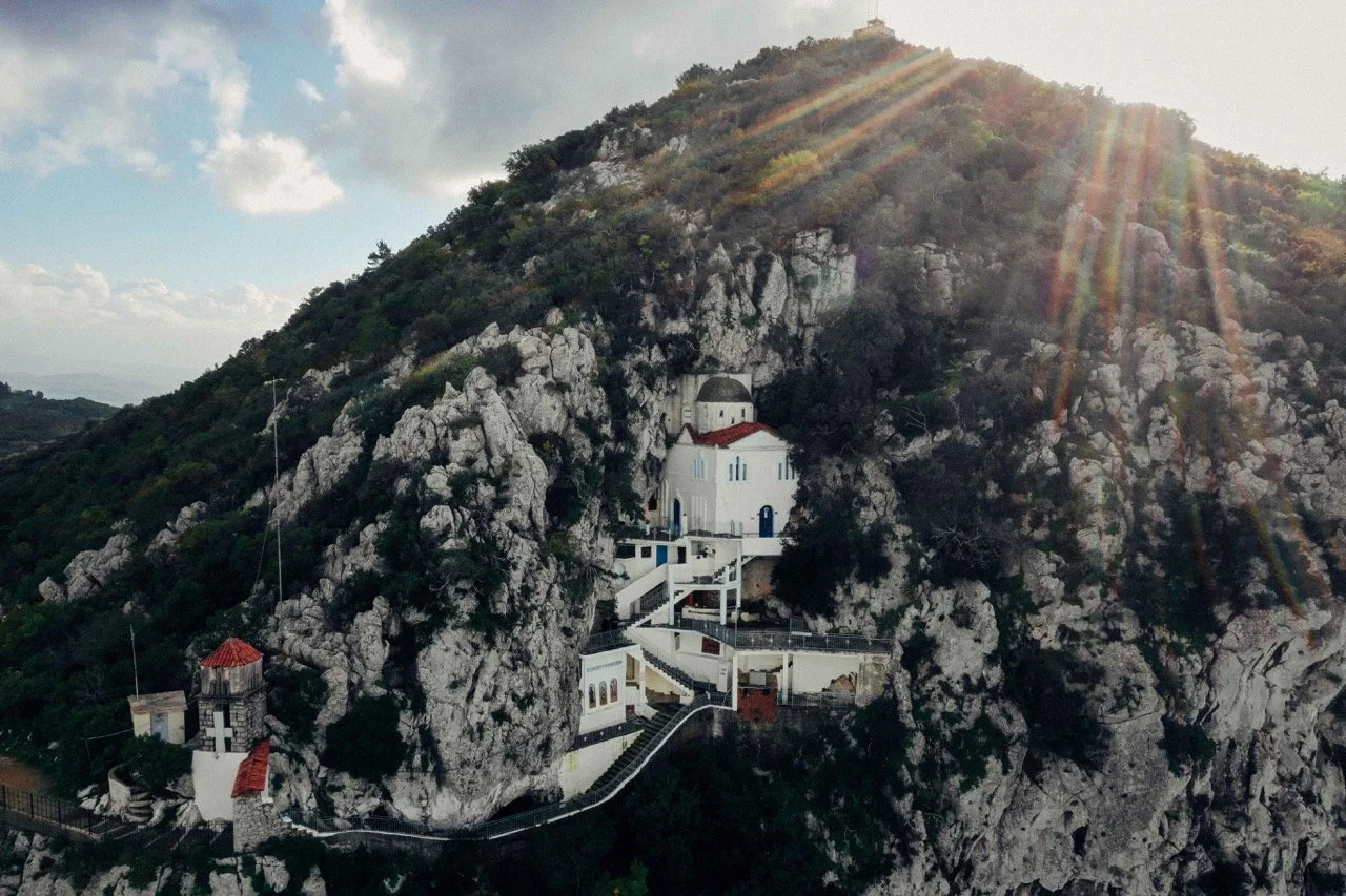 A church built into a rocky hillside with stairs leading up to it, surrounded by greenery, under sunlight with rays shining down.