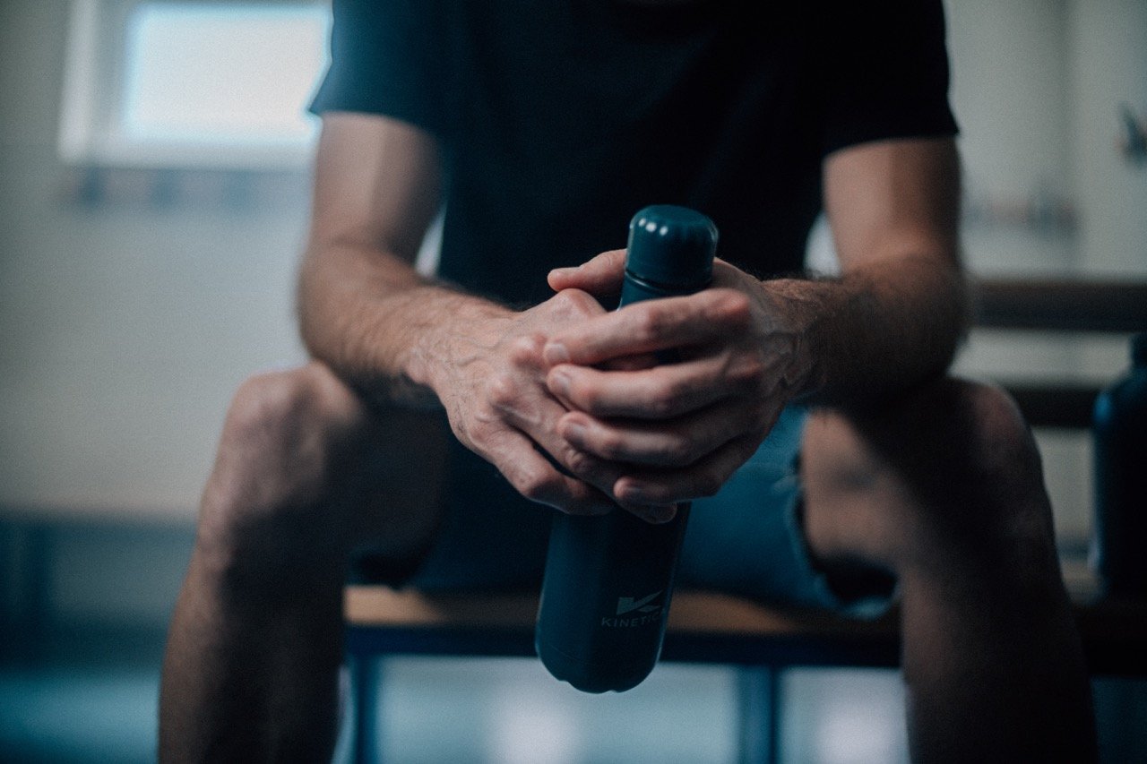 A man sitting on a bench in a gym, holding a water bottle with both hands.