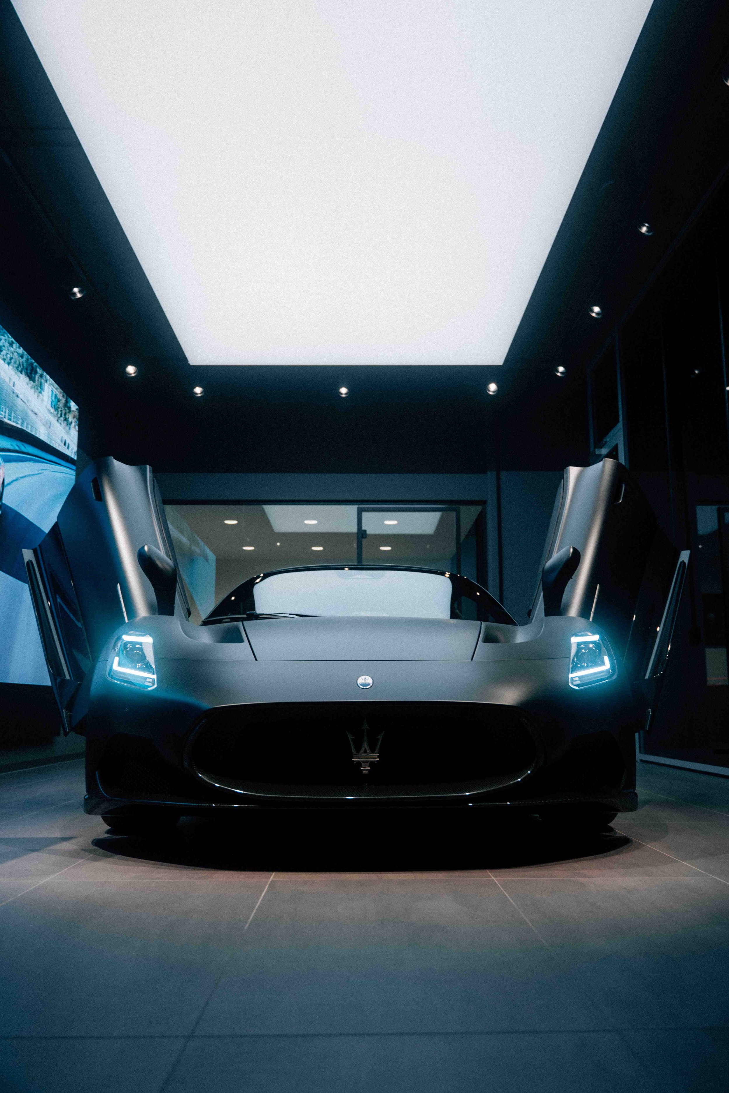 A sleek, black luxury sports car with its gull-wing doors open, displayed in a modern showroom with a large illuminated ceiling panel.